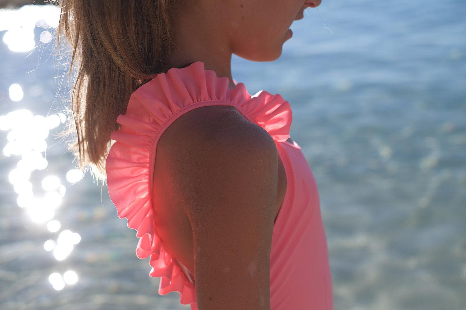 Girl in pink ruffle swimsuit by the water, sunlight reflecting off the sea