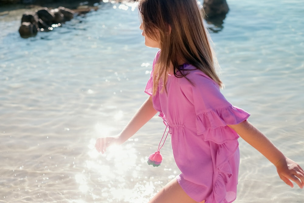 Young girl in pink dress playing near clear sea water with sunlight reflections on beach