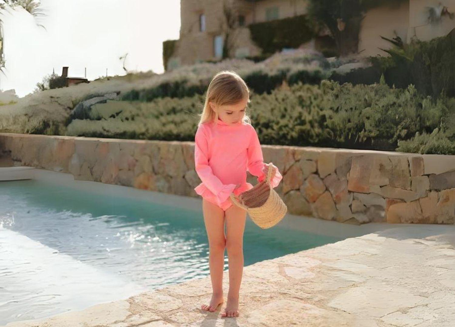 Girl in long-sleeve pink swimsuit by pool holding a straw bag, sun protection beachwear
