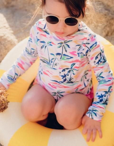 Child in colorful long-sleeve UV swimsuit and sunglasses sitting on yellow float at the beach