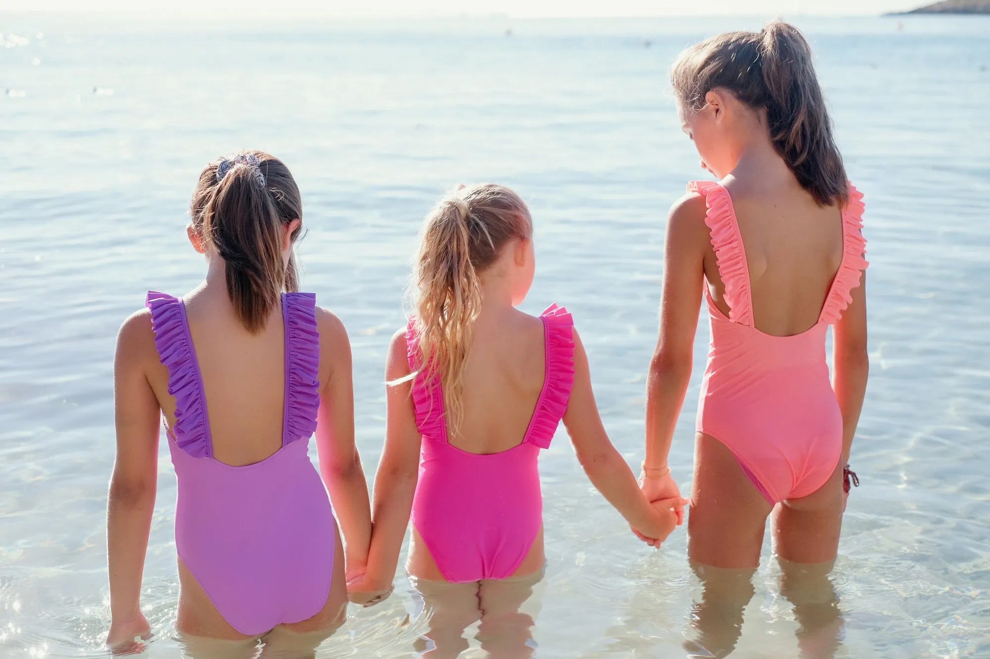 Three girls in ruffled anti-UV swimsuits standing in shallow beach water, backs to camera.