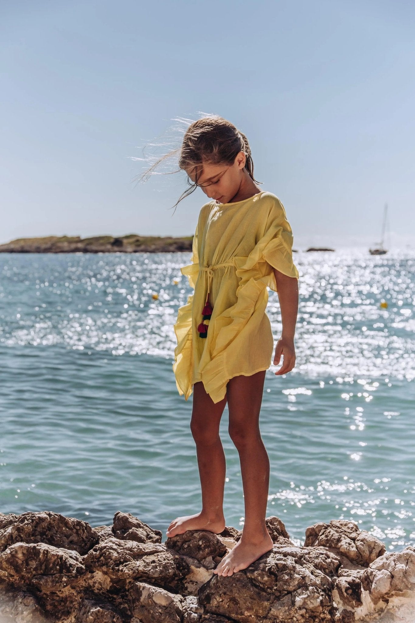 Girl in a yellow caftan dress standing on rocks by the seaside, sunny beachwear scene