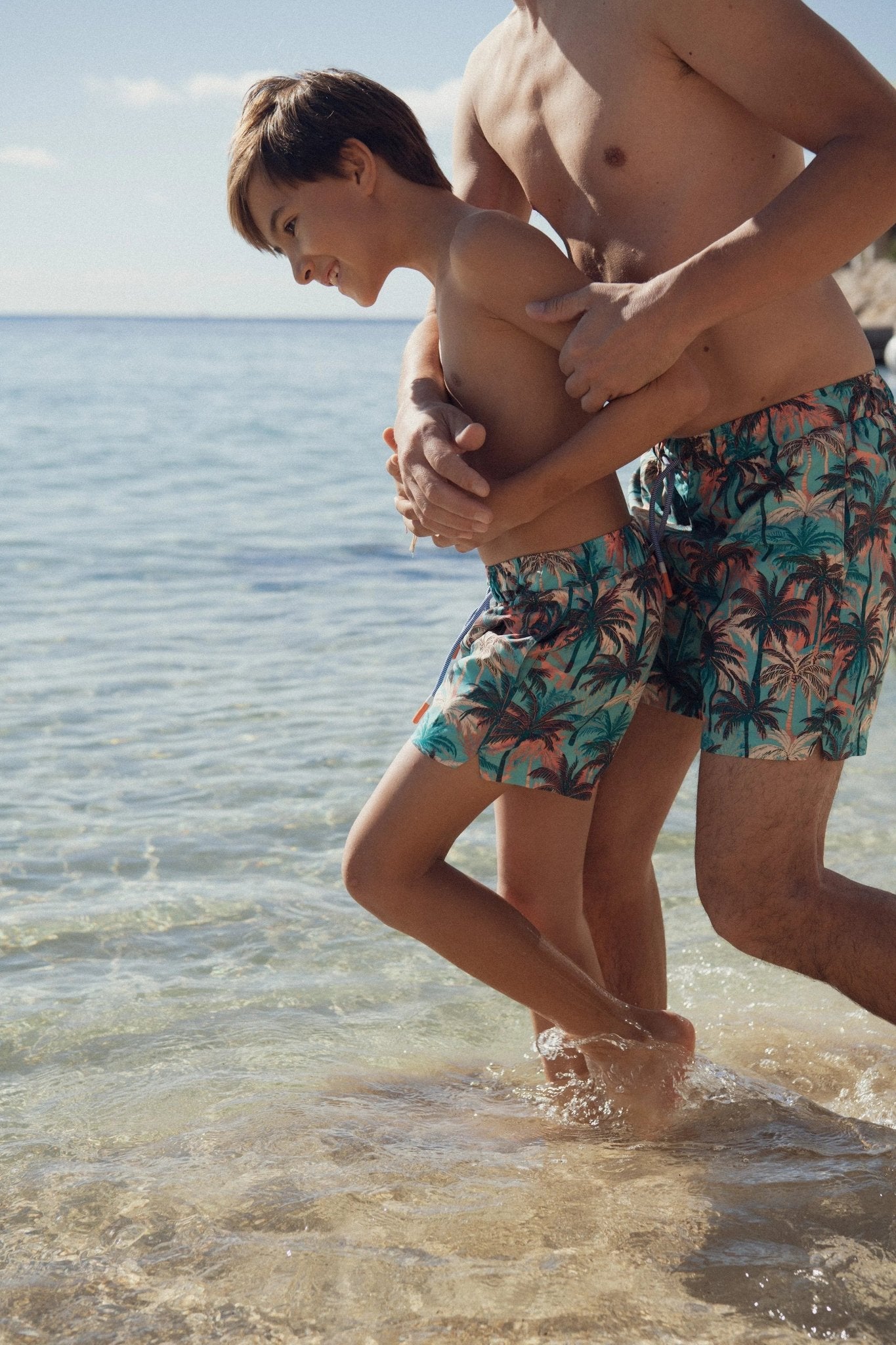 Boy and man wearing matching tropical swim shorts playing at the beach shoreline