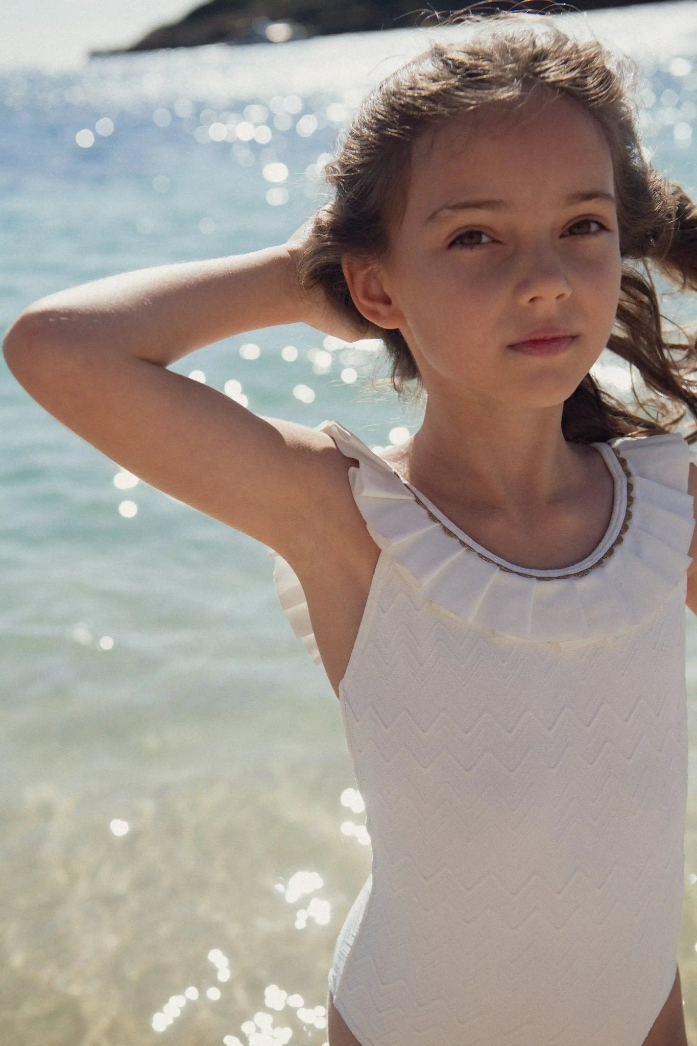 Girl in white ruffled one-piece swimsuit standing by the sea on a sunny day