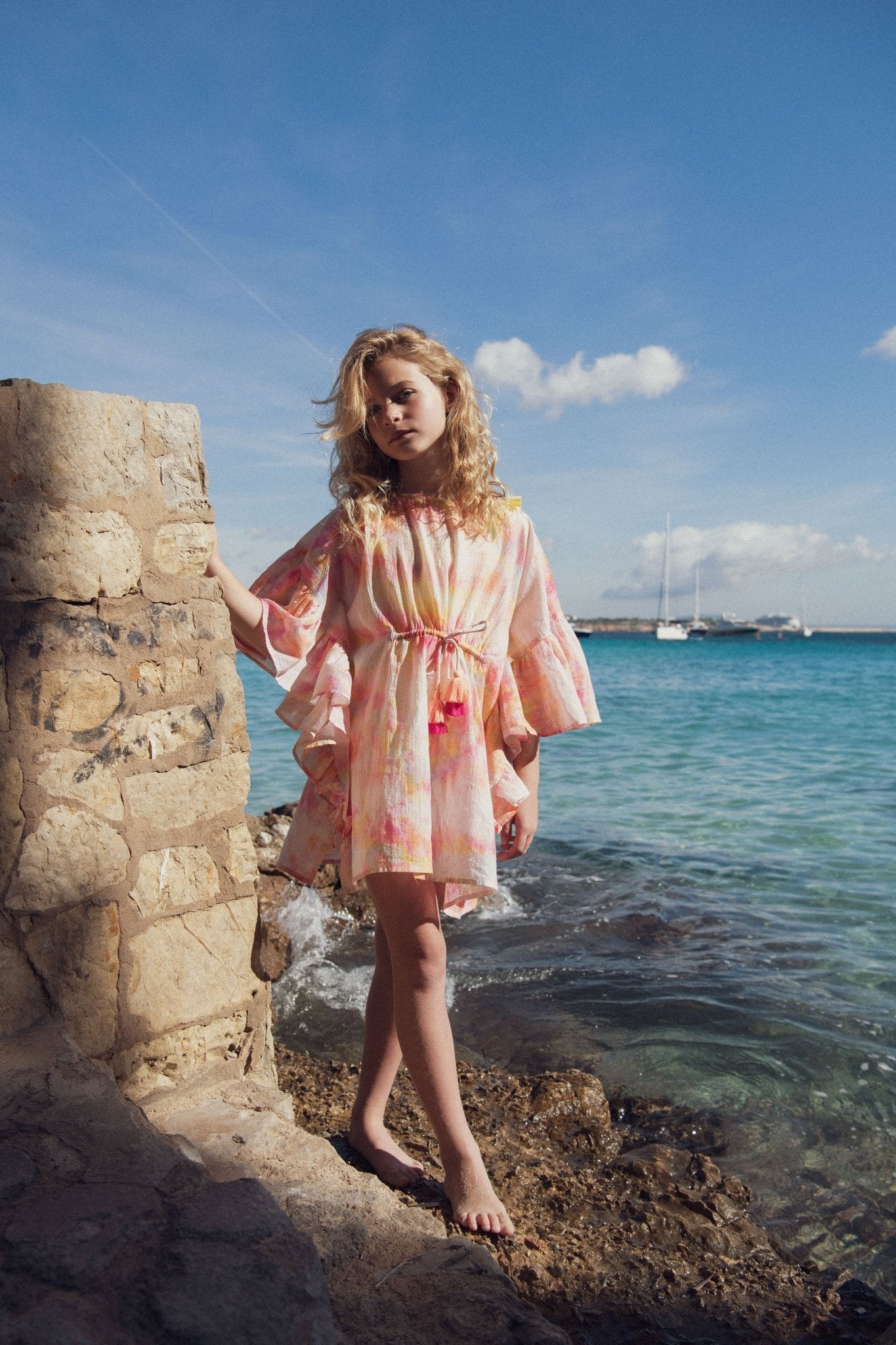Girl in pastel beach kaftan standing by stone wall near the sea under a blue sky