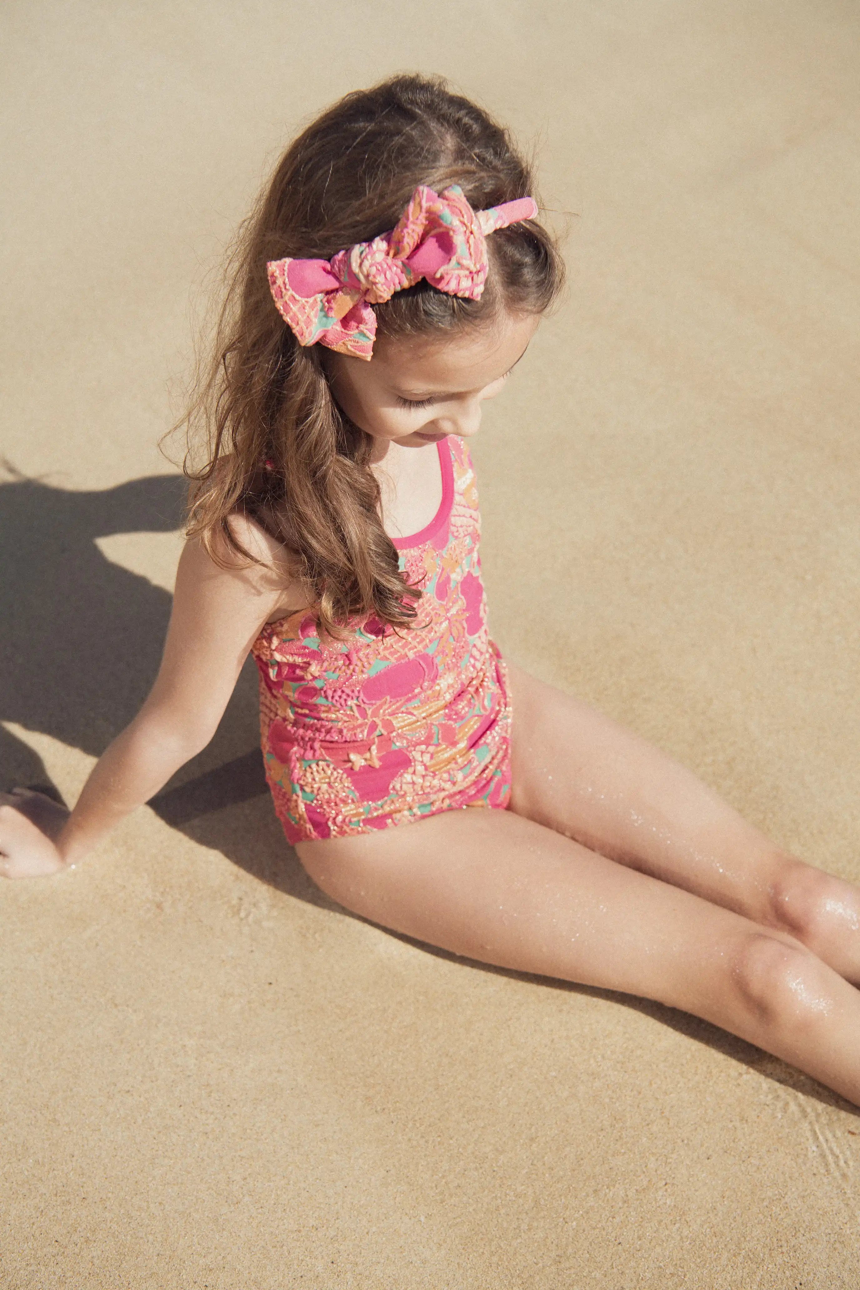 Girl in pink Lison Paris UV protection swimsuit and matching headband sitting on sandy beach