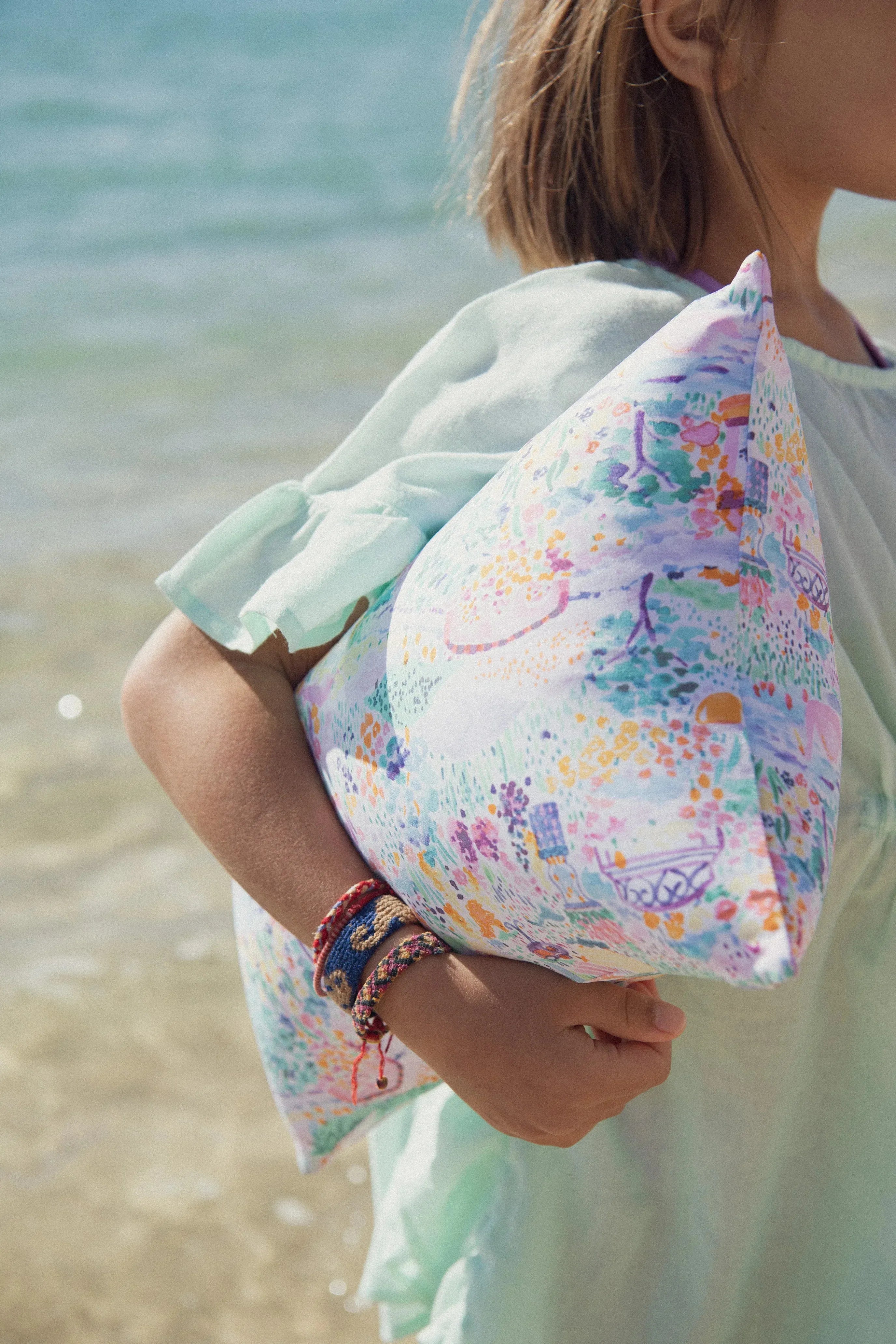 Child in pastel beachwear holding a colorful cushion by the sea, Lison Paris summer style