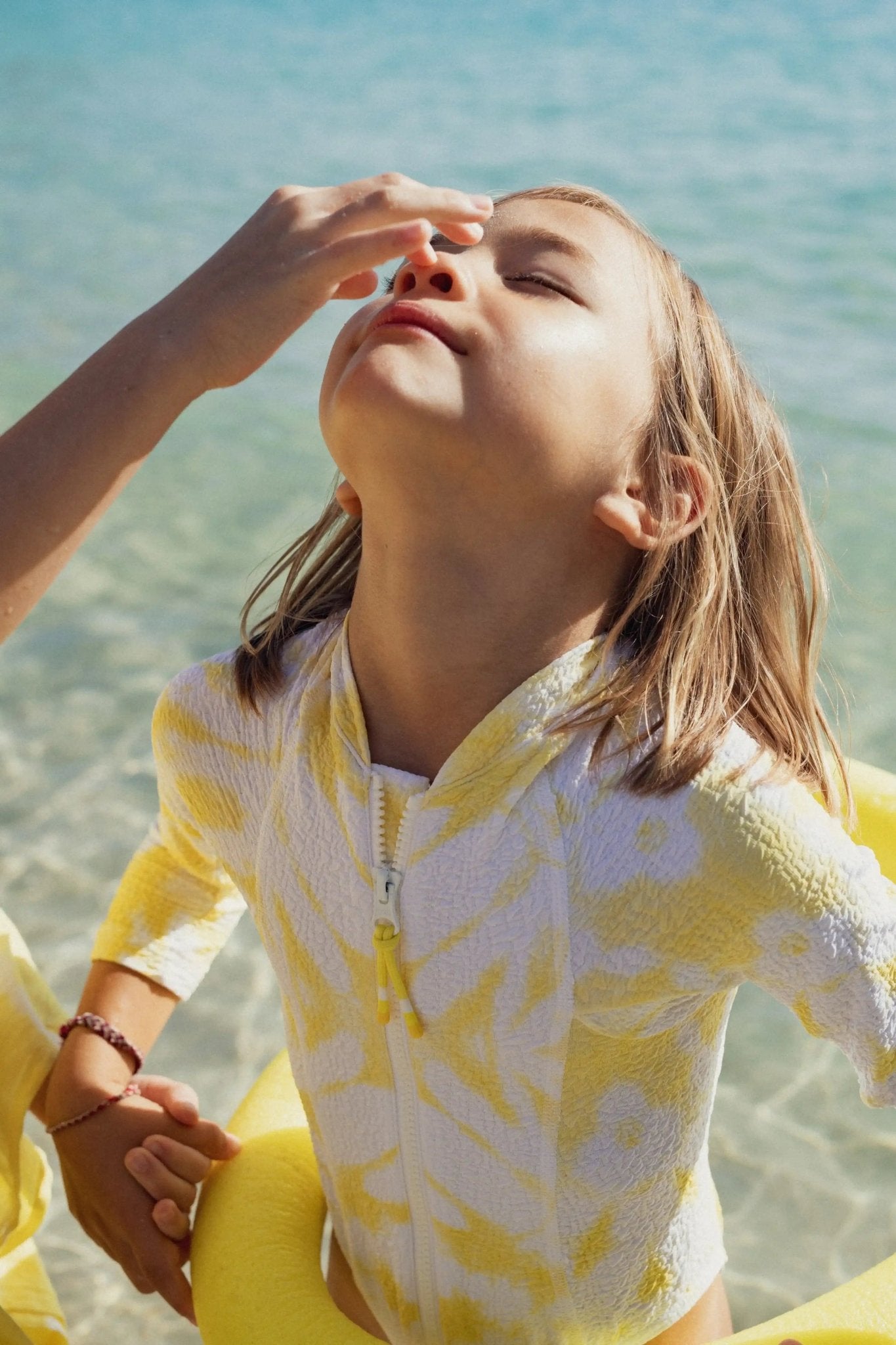 Child in yellow and white UV swimwear at the beach, applying sunscreen by the sea