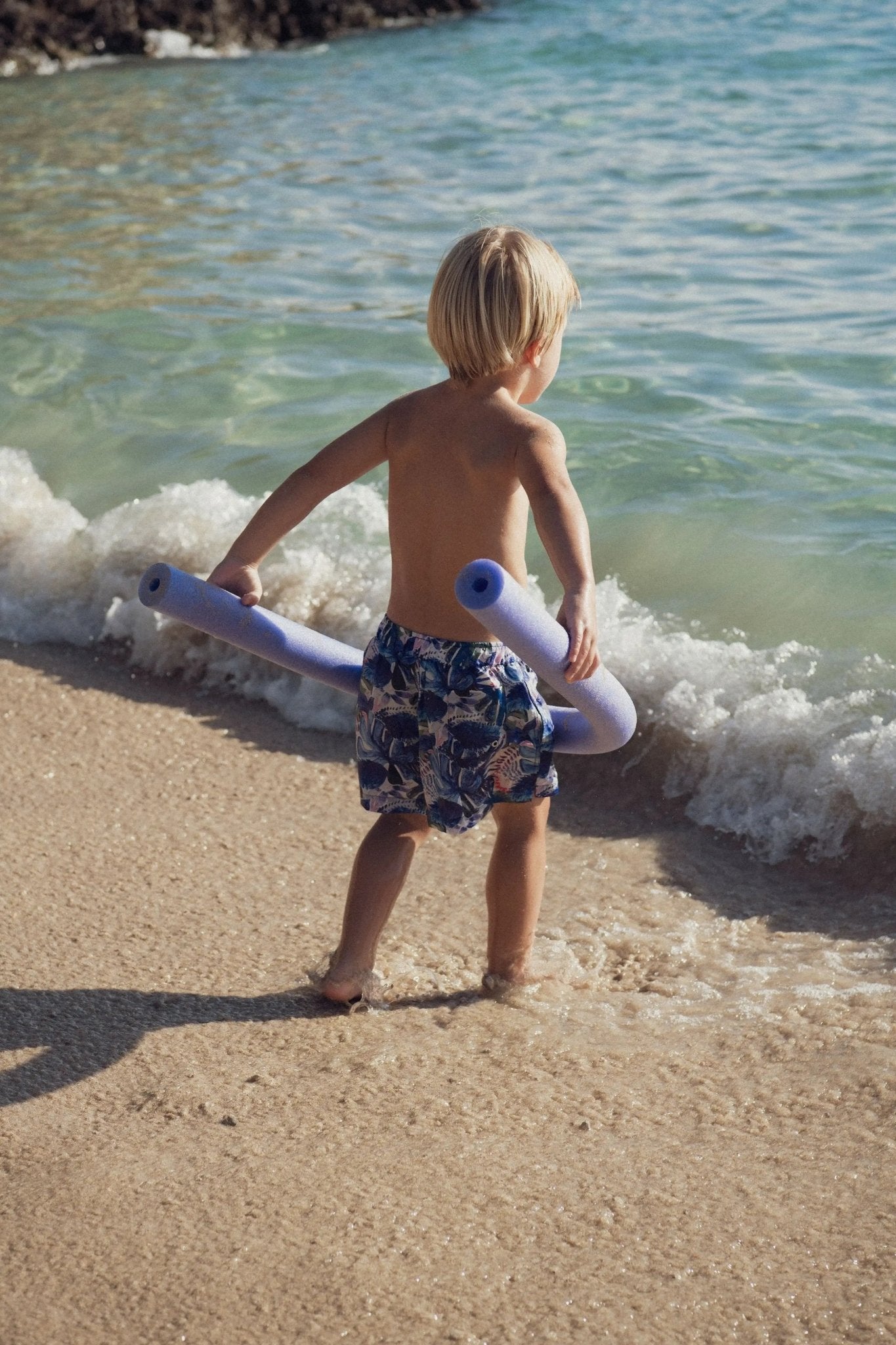 Little boy in patterned swim trunks holding pool noodles at the beach shoreline