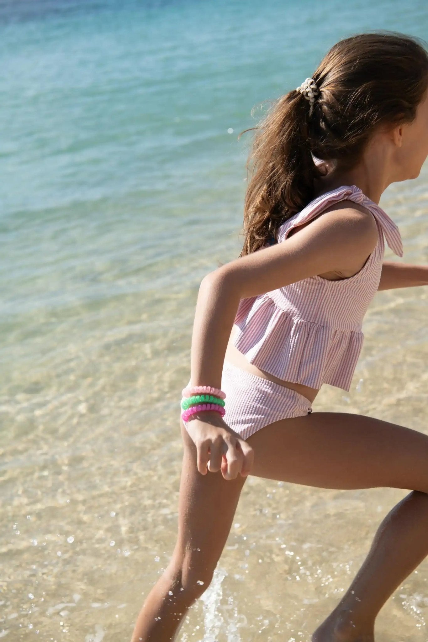 Girl in pink ruffled swimsuit playing by the sea, wearing colorful bracelets.