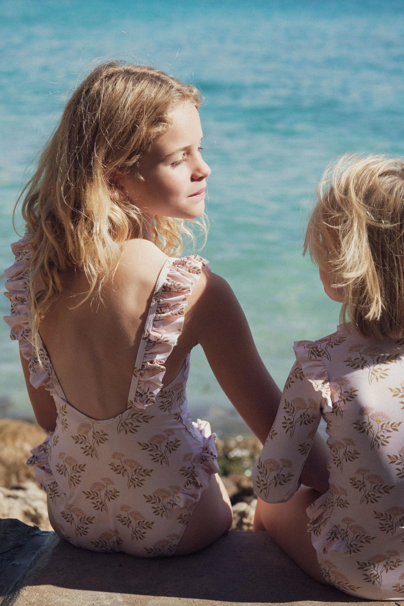 Two girls in floral ruffle swimsuits sitting by the sea on a sunny day