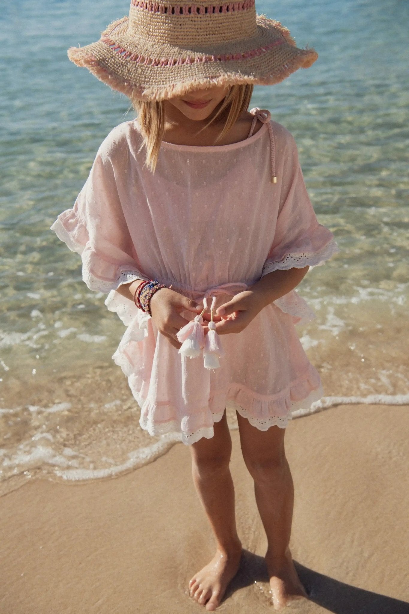 Girl in pink caftan dress and straw hat standing barefoot on the beach near the water