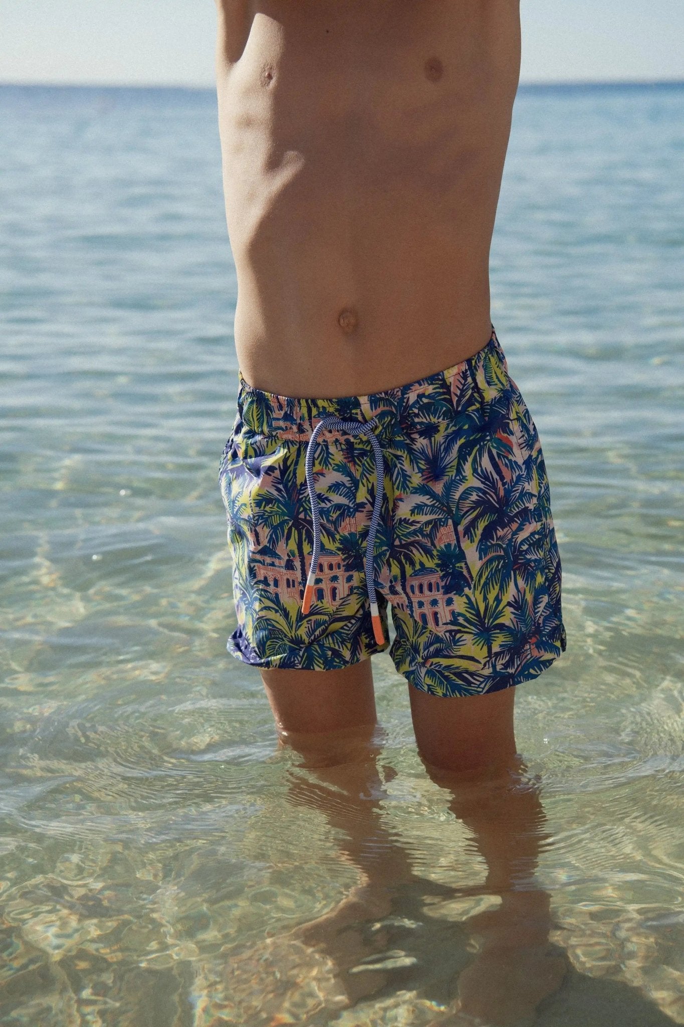Boy wearing blue jungle print swim shorts standing in clear shallow sea water