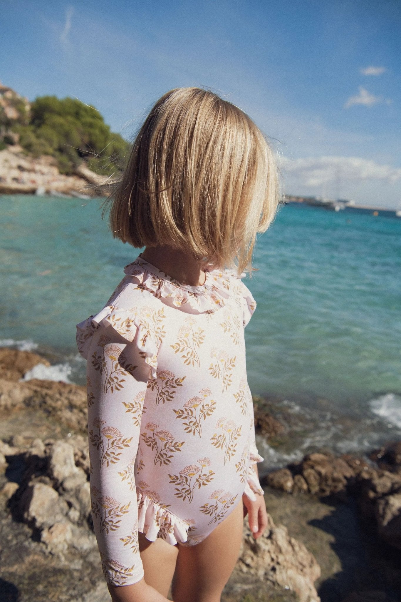 Girl in long-sleeve floral swimsuit on rocky beach by the sea under sunny sky