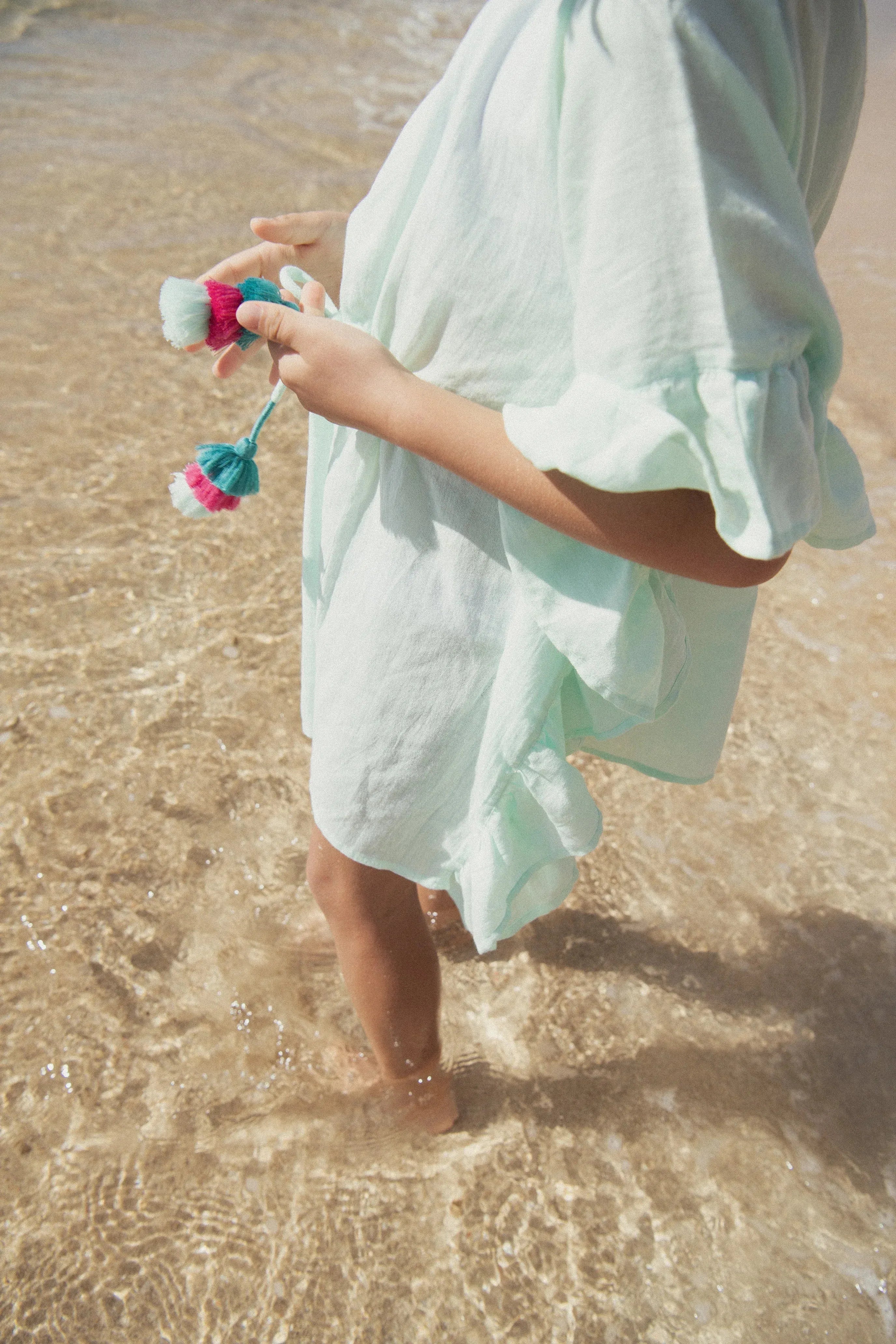 Child in aqua beachwear by Lison Paris standing in shallow water, holding colorful tassels