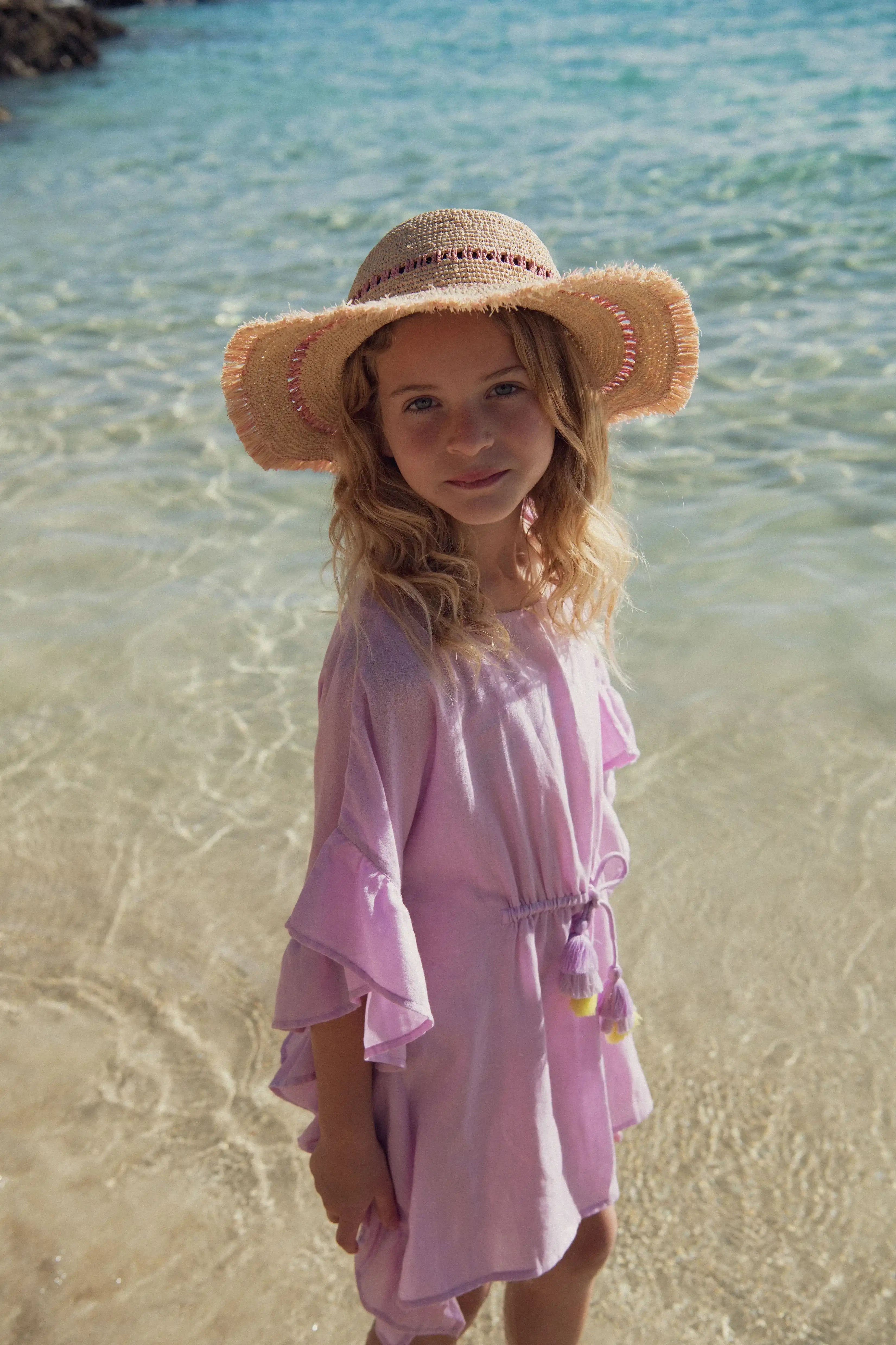 Girl in lilac beach kaftan and straw hat standing on the shore, Lison Paris beachwear collection.