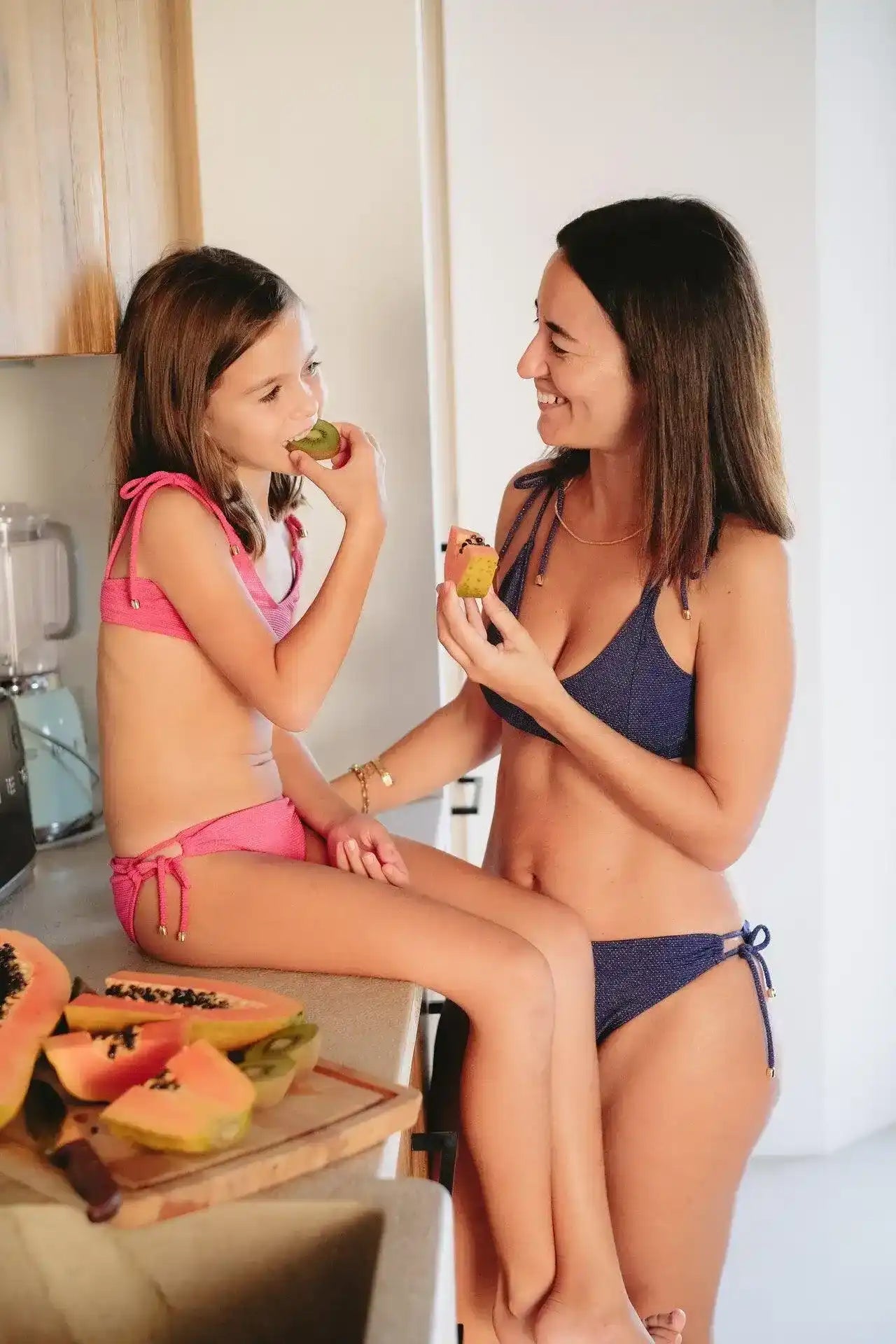 Girl in pink textured bikini eating fruit with woman in blue bikini in a bright kitchen