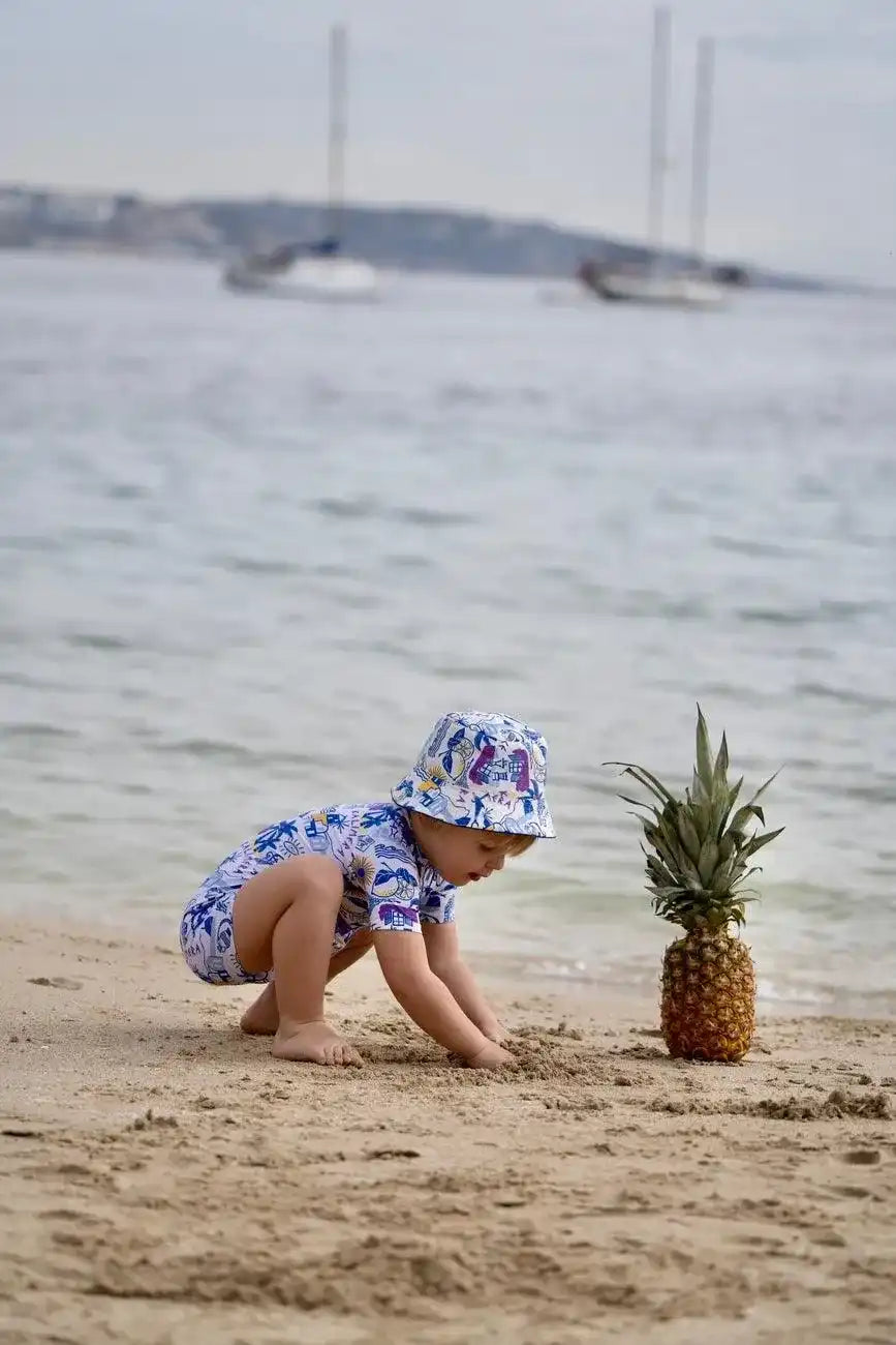 Toddler in Lison Paris anti-UV swimsuit and hat playing on sandy beach near pineapple