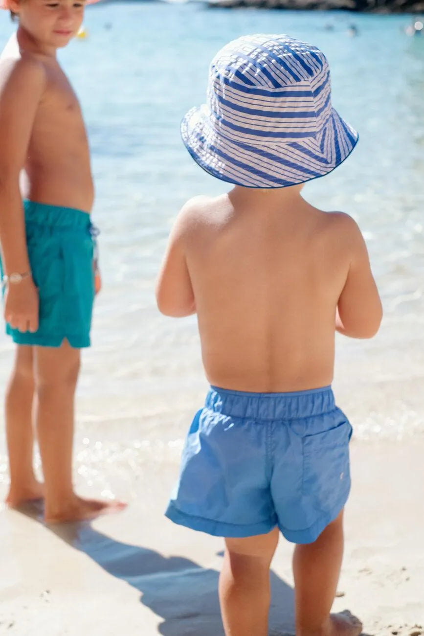Two young boys at the beach, one wearing blue striped sun hat and shorts, the other in turquoise swim trunks