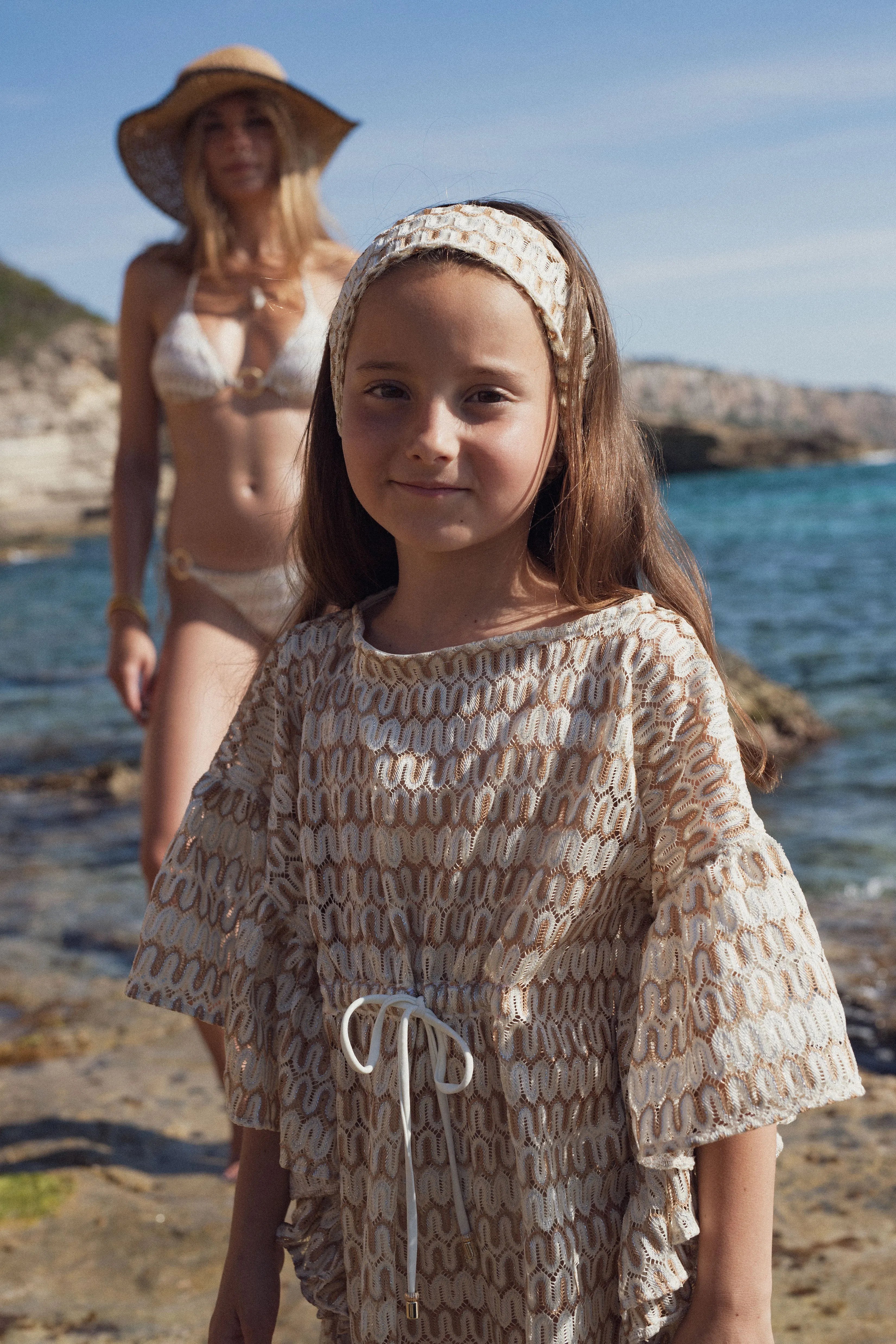 Young girl in beige and white crochet dress and headband on rocky beach with woman in bikini and sunhat