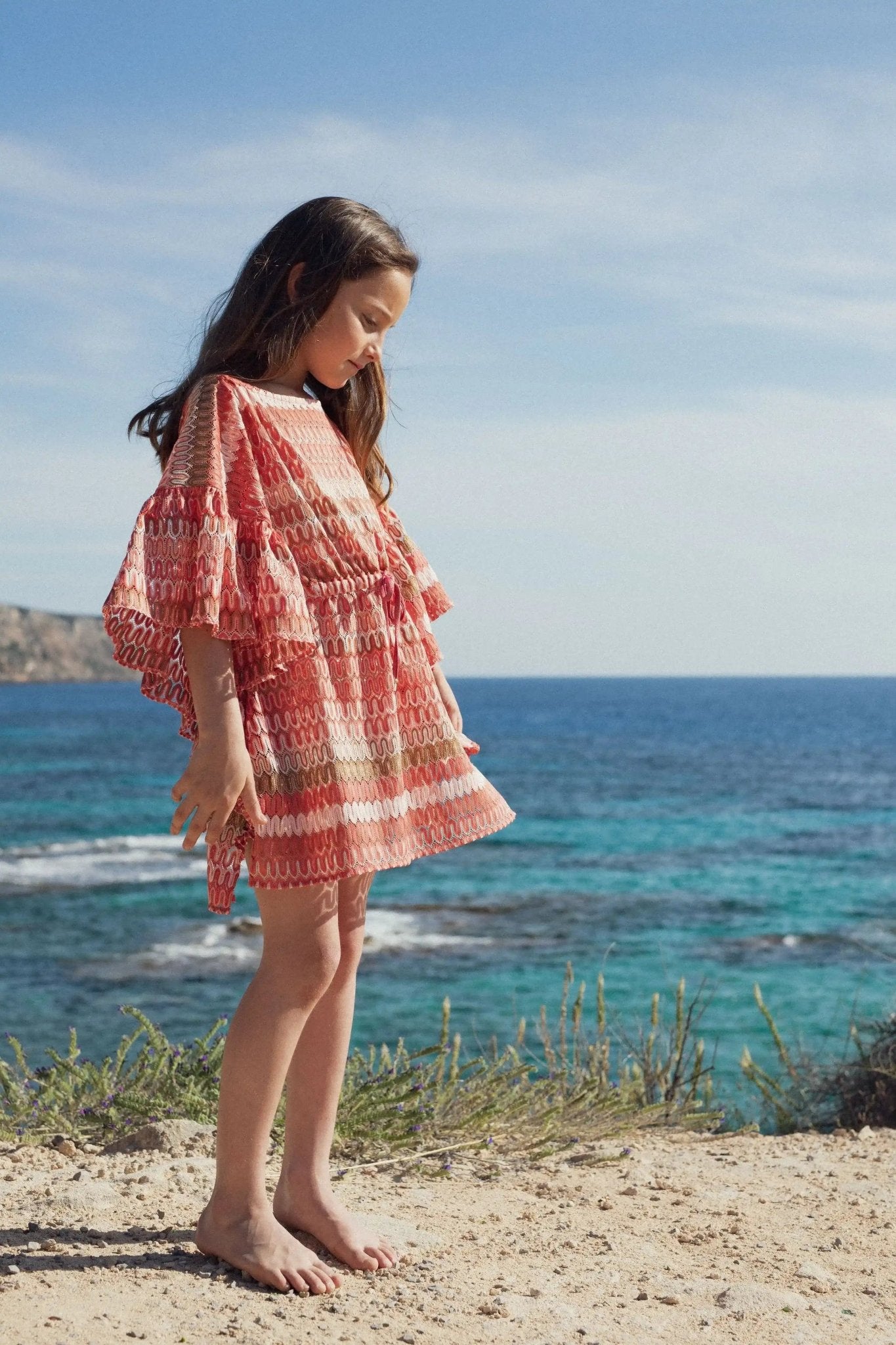 Girl in coral and gold crochet caftan standing barefoot by the sea on a sunny day
