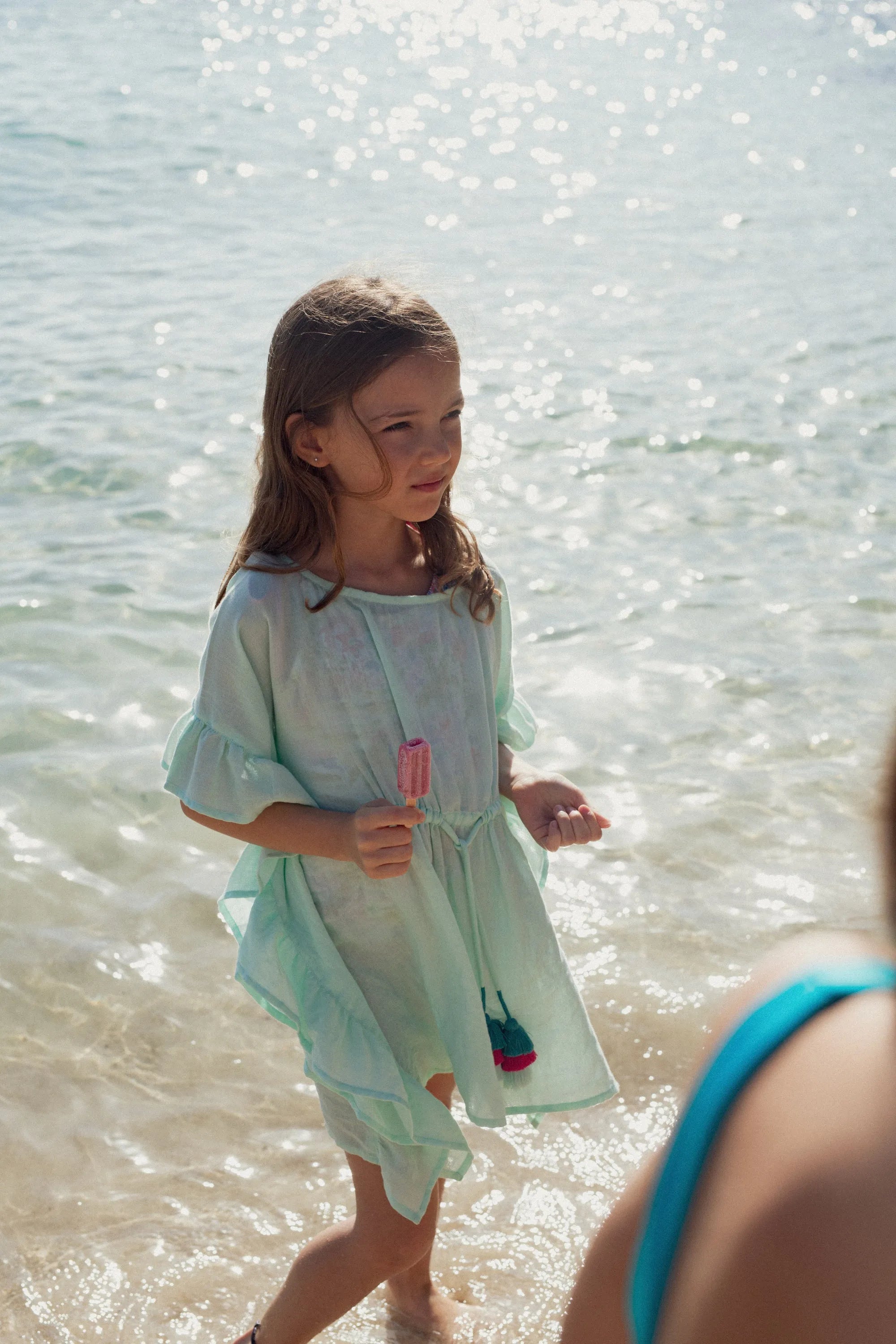 Young girl in aqua caftan with pompons holding pink popsicle by the sea shore