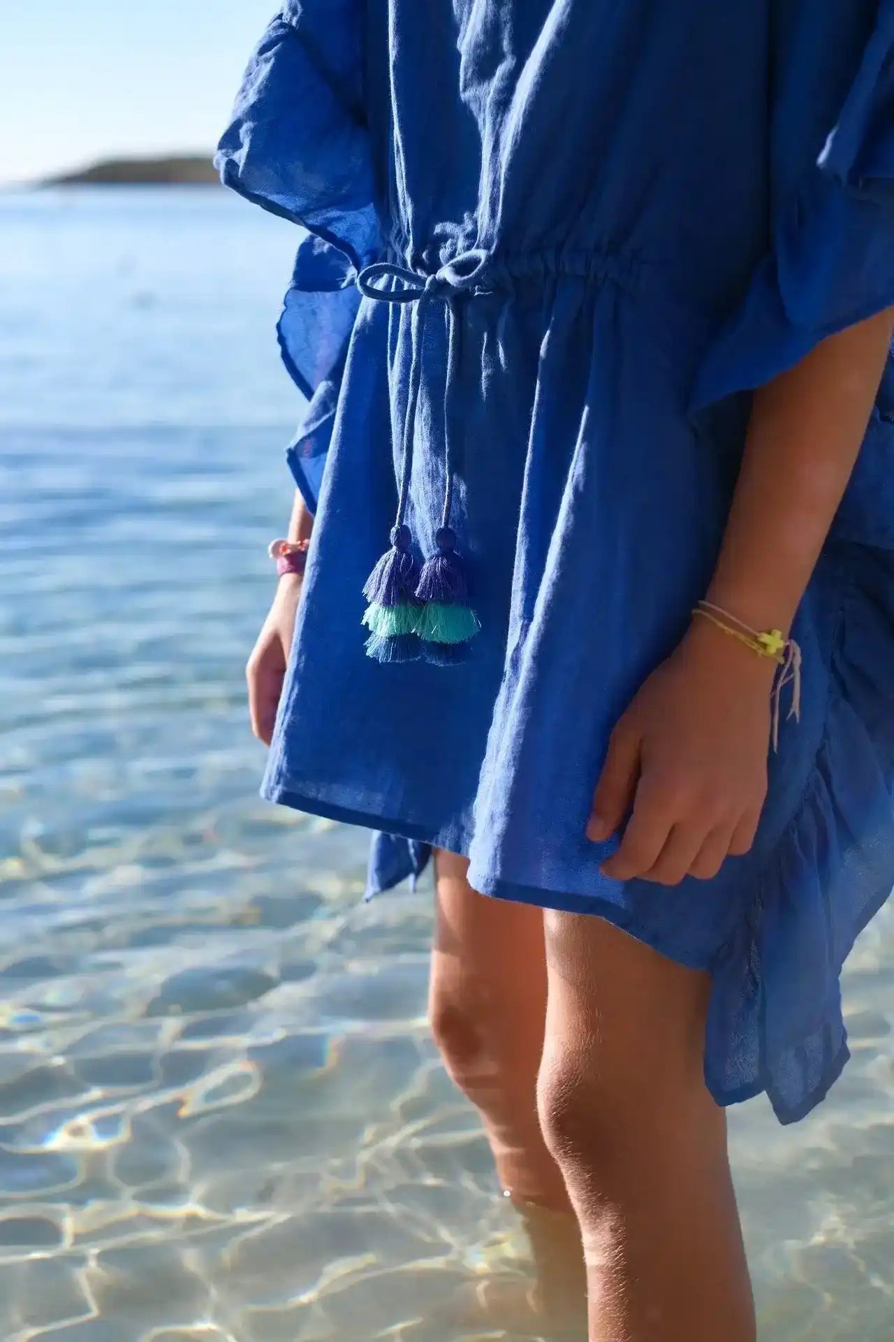 Girl wearing blue caftan with pompom details standing in shallow clear water