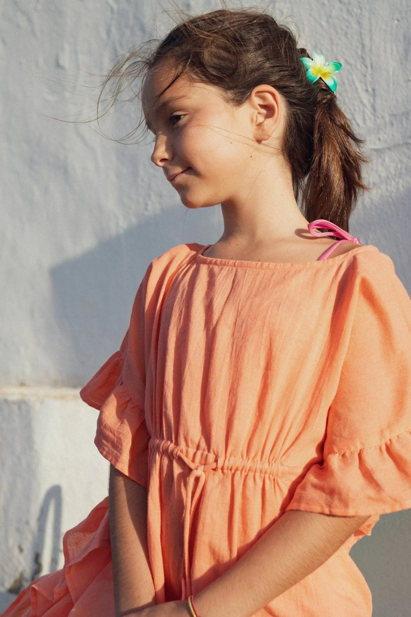 Girl in coral pompom caftan with ruffled sleeves and flower hair tie outdoors
