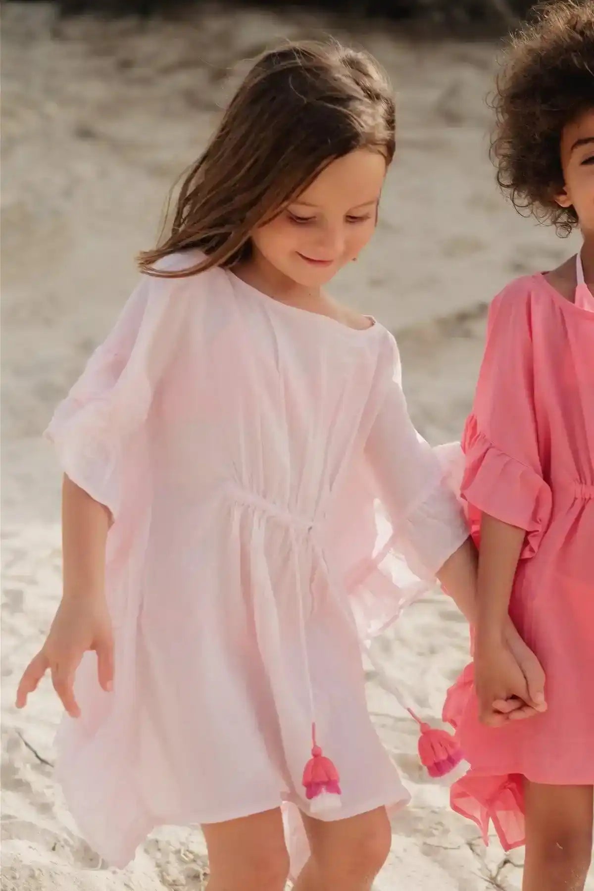 Girl in pale pink caftan dress with pompom details on the beach, holding hands