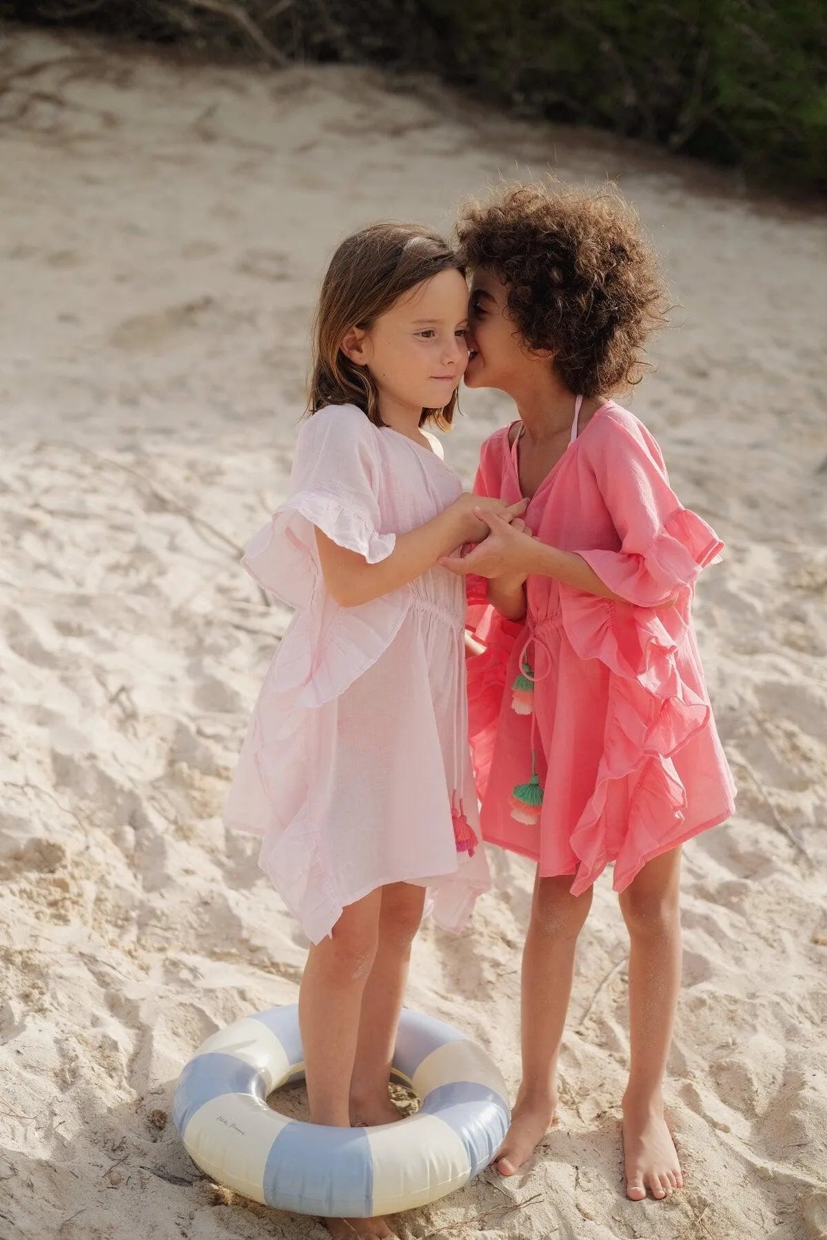 Two young girls on sandy beach wearing pink ruffled caftans, one whispering to the other