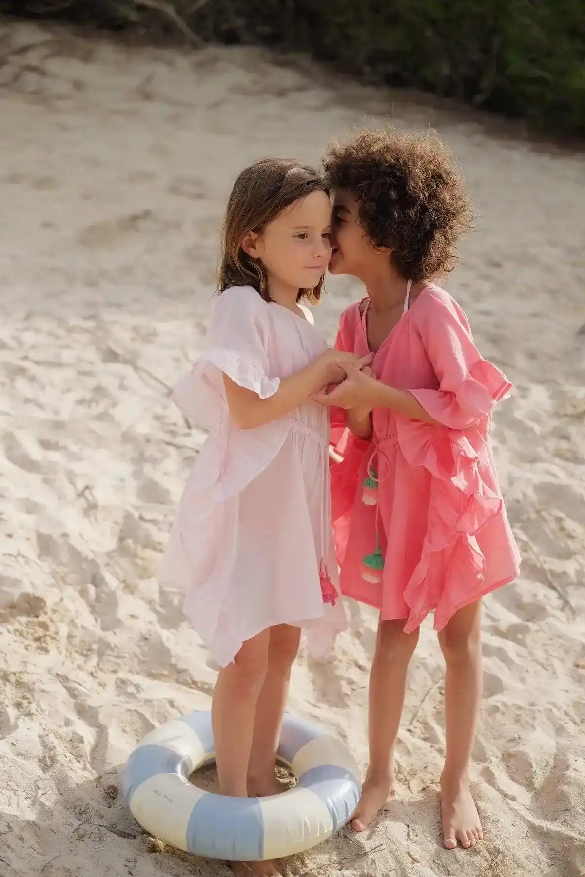 Two girls in pink caftans with pompons standing on sandy beach, one whispering to the other