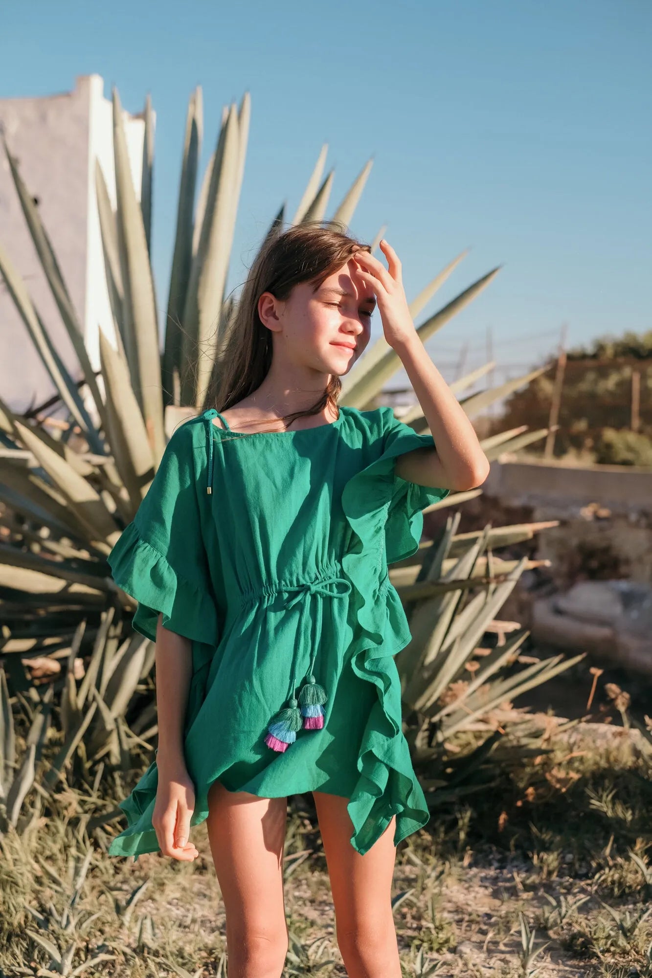 Girl wearing green ruffled caftan dress with tassels standing outdoors near large agave plants