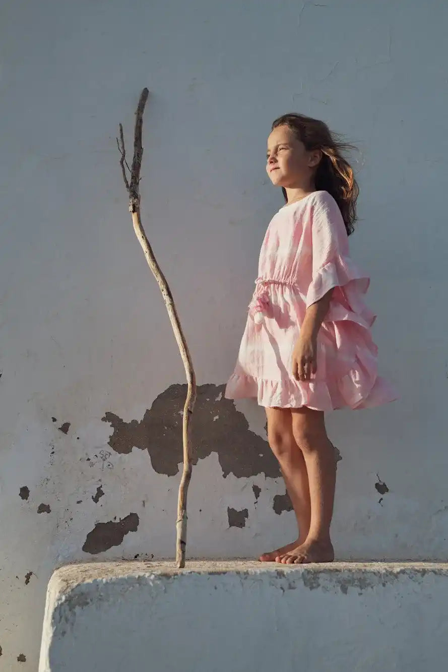 Girl in pink tie-dye caftan from Lison Paris standing barefoot outdoors by a white wall