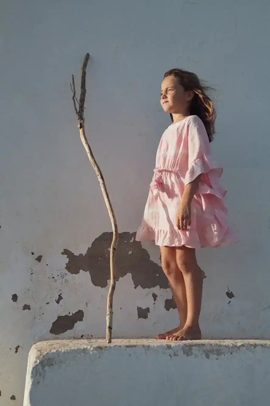 Girl in light pink tie-dye caftan dress standing barefoot outdoors by a white wall