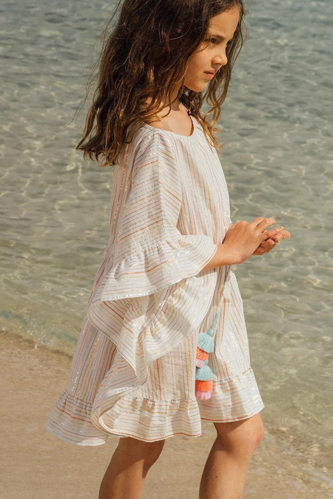 Young girl wearing white striped caftan with colorful pompons standing on beach near clear water