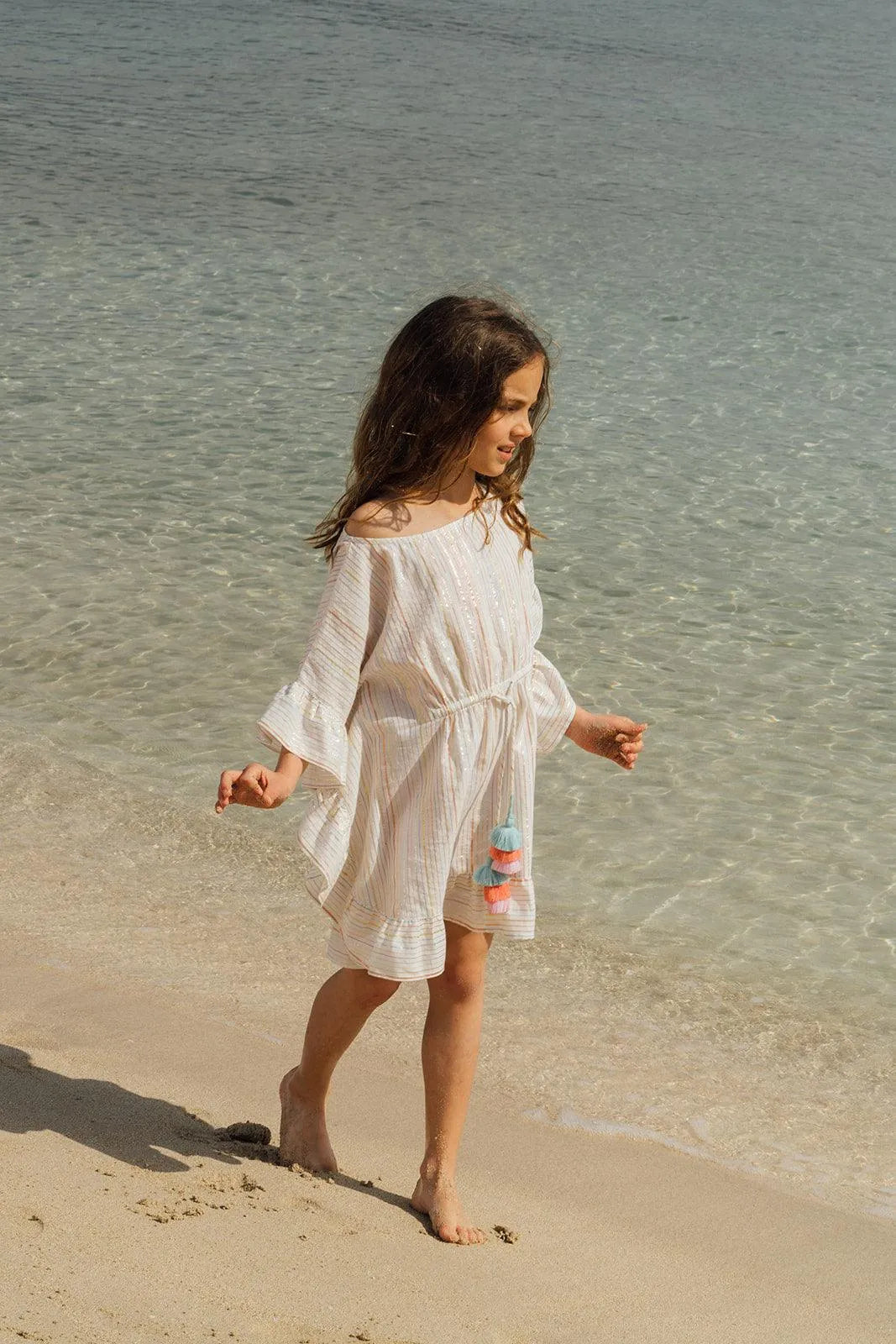 Young girl walking barefoot on the sandy beach wearing a light striped dress with colorful pompons