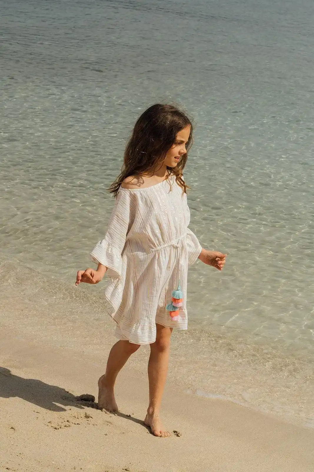 Girl in Lison Paris striped caftan with pompons walking on the beach by clear water