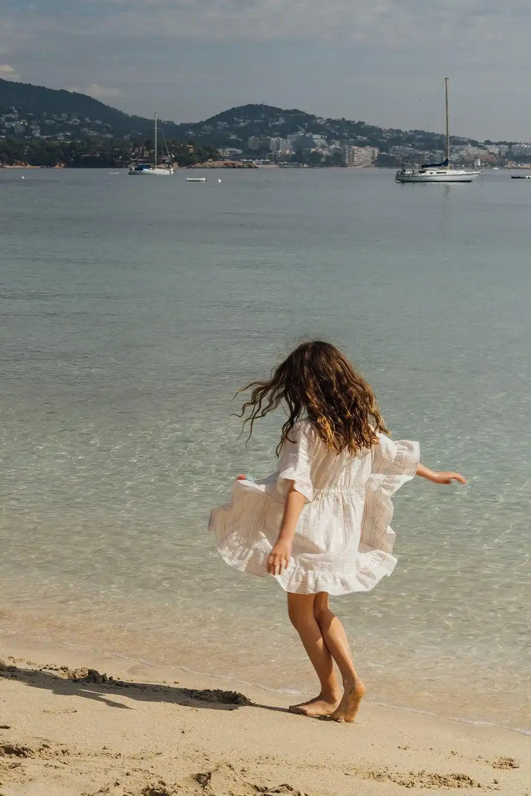 Girl in a white Lison Paris beach dress twirls on sandy shore by calm sea, sailboats in background