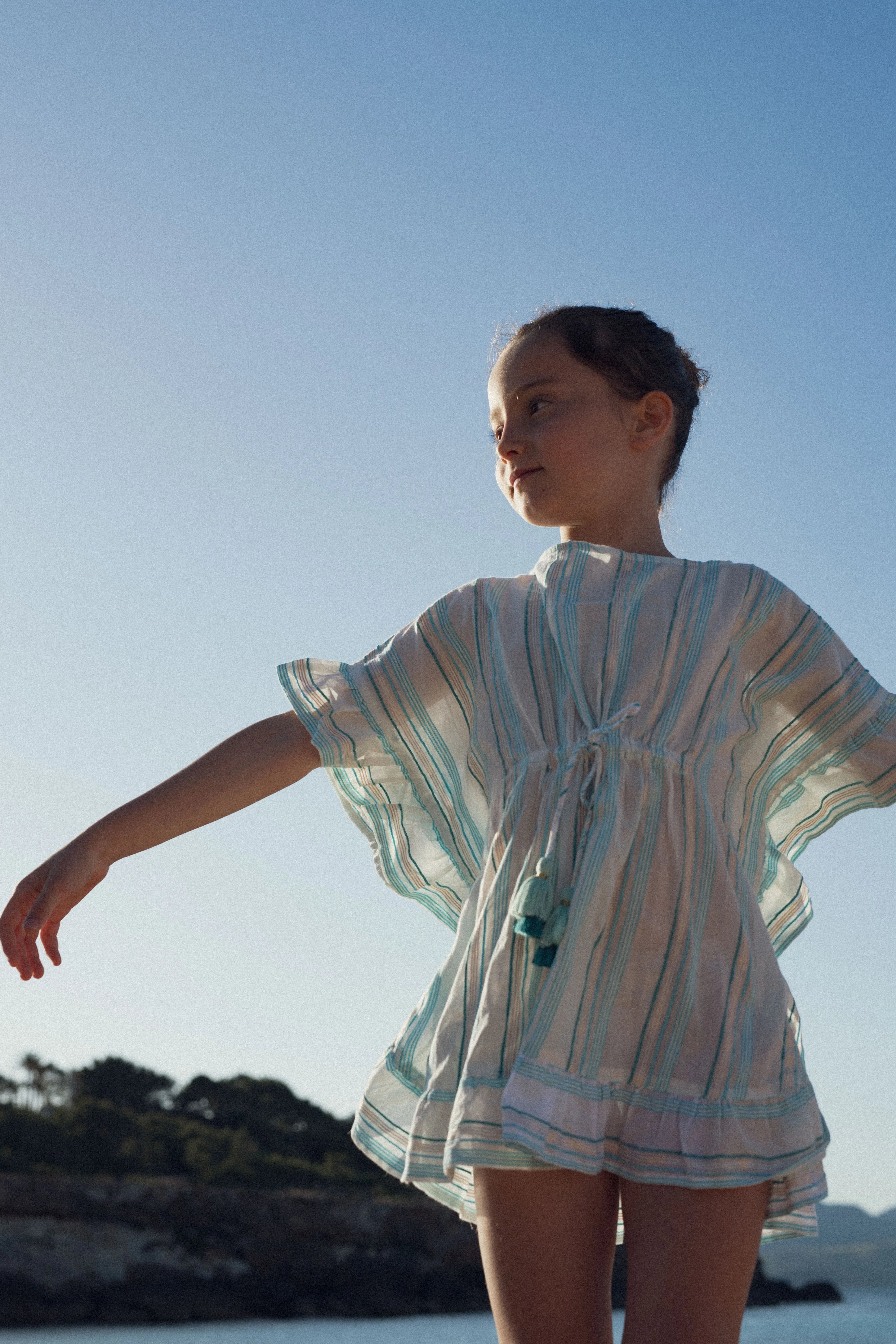 Young girl in a blue-striped beach caftan dress standing by the seaside at sunset