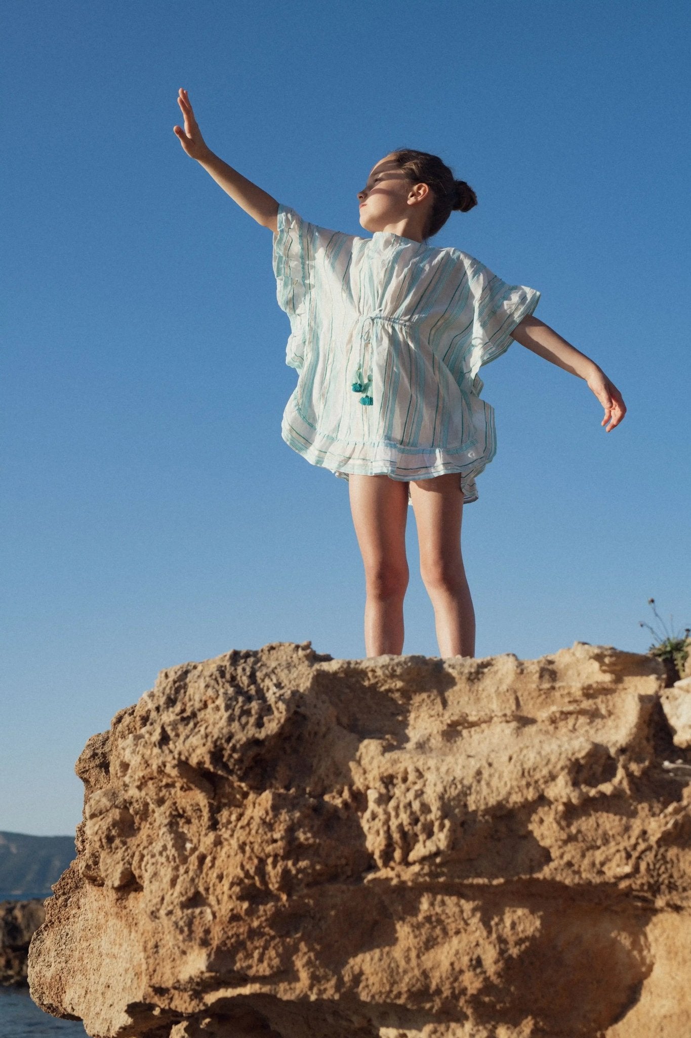 Girl in aqua and gold striped caftan beachwear standing on a rock under blue sky