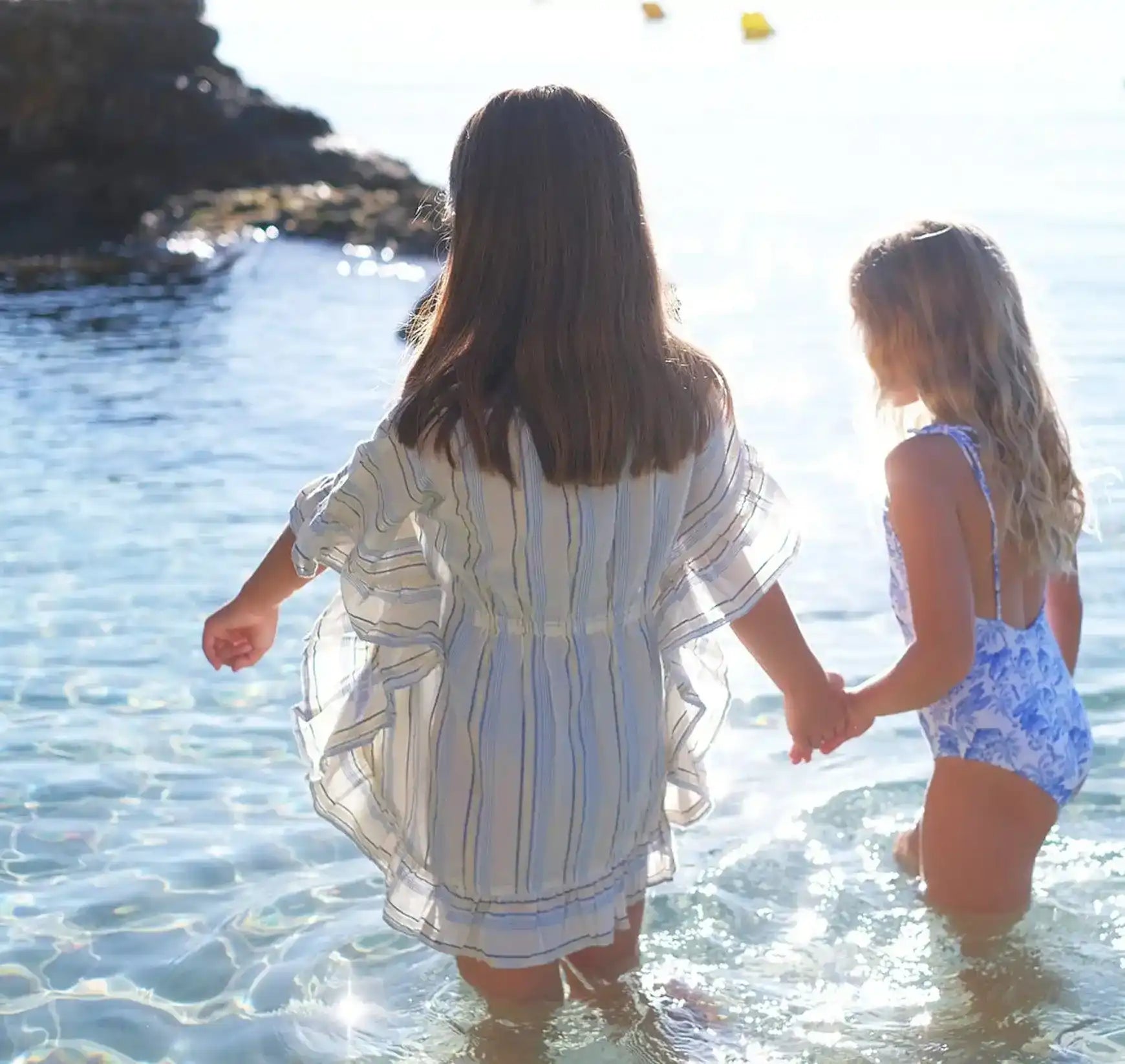 Two girls in striped caftan and blue floral swimsuit holding hands in shallow beach water