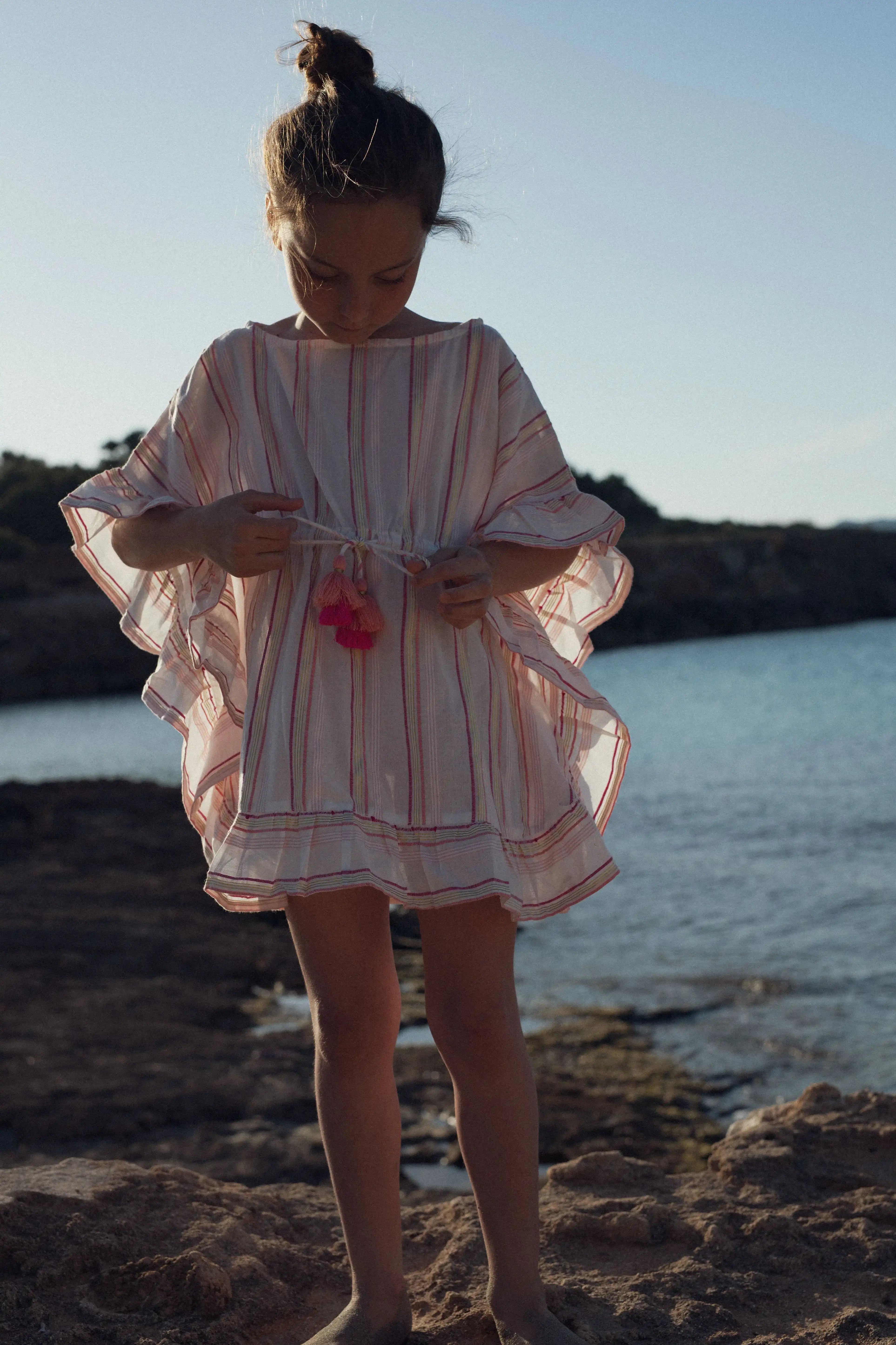 Girl in pink striped Lison Paris caftan with tassels by the seaside, beachwear for kids
