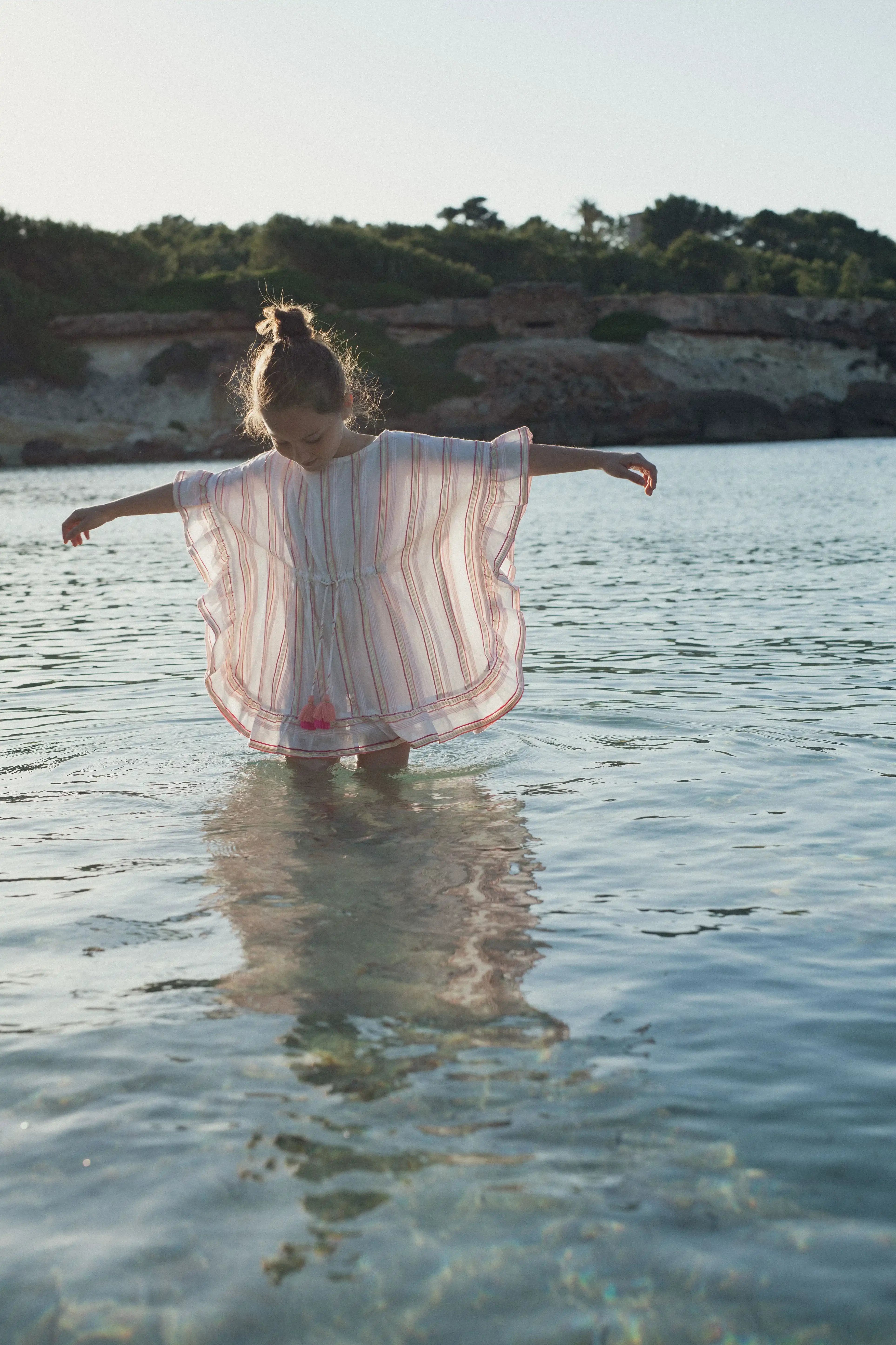 Girl wearing pink and gold striped caftan stands in shallow water, Lison Paris beachwear