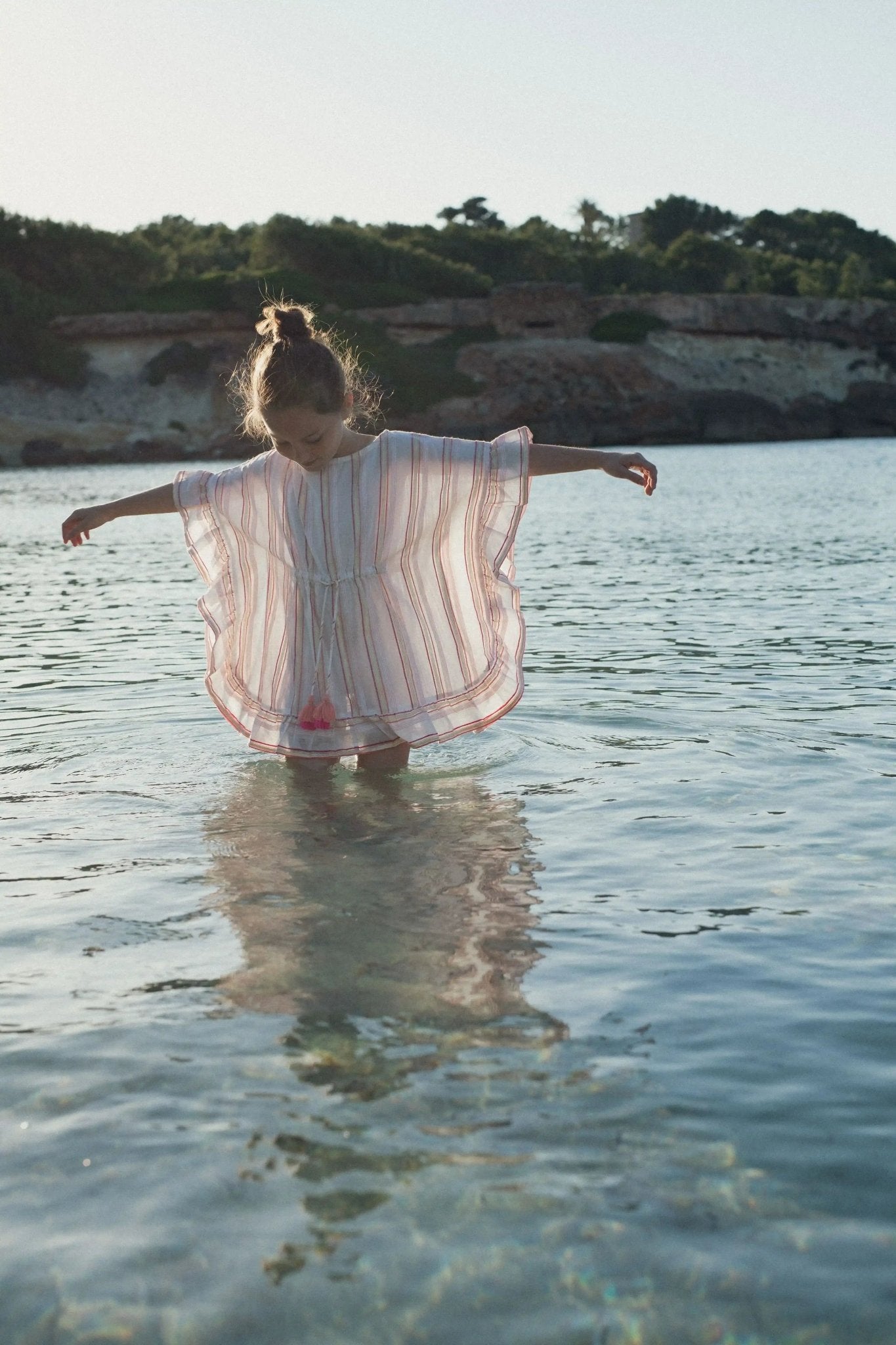 Girl in striped pink and gold caftan standing in clear shallow water by the beach
