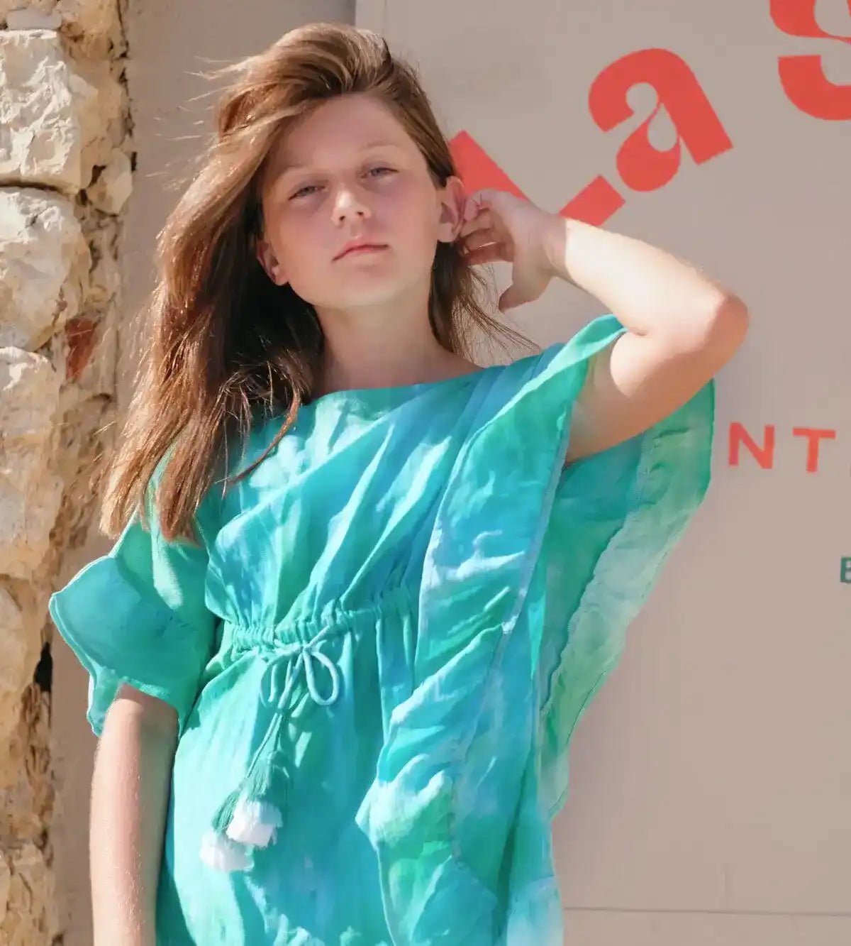 Girl in green and blue tie-dye caftan, posing outside near stone wall in sunlight