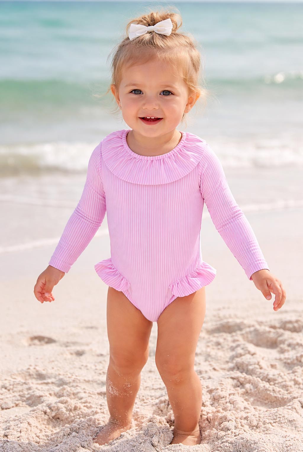Toddler girl in long sleeve pink ruffled swimsuit standing on beach sand by the sea