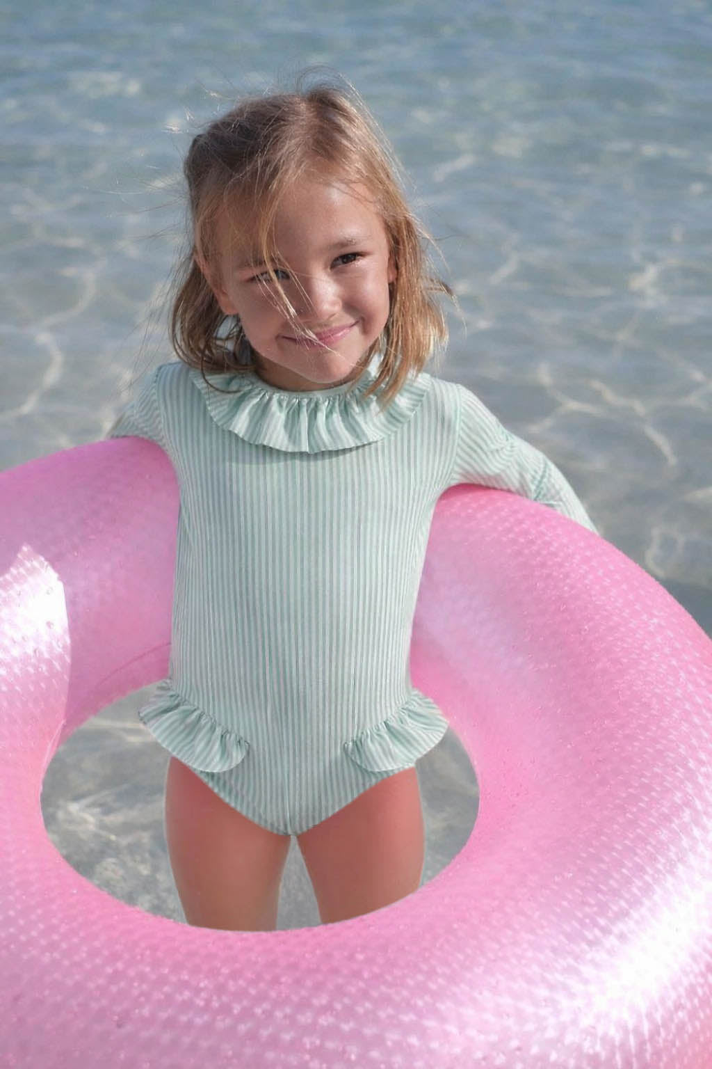 Girl in long-sleeve striped swimsuit with ruffles, holding pink float, standing in shallow water.