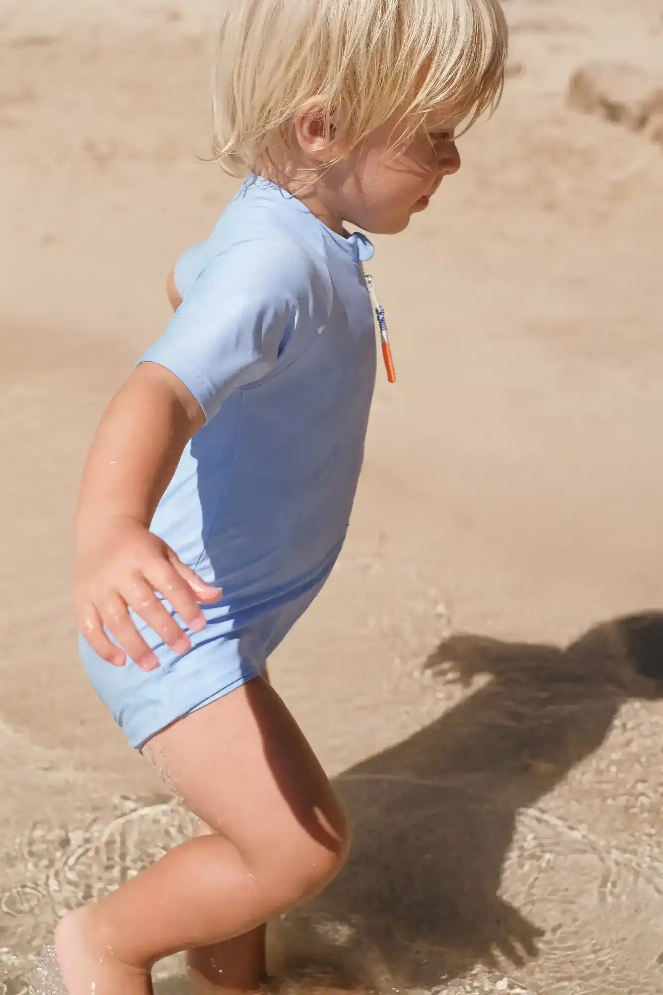 Toddler boy wearing light blue short-sleeve UV-protection swimsuit playing on sandy beach