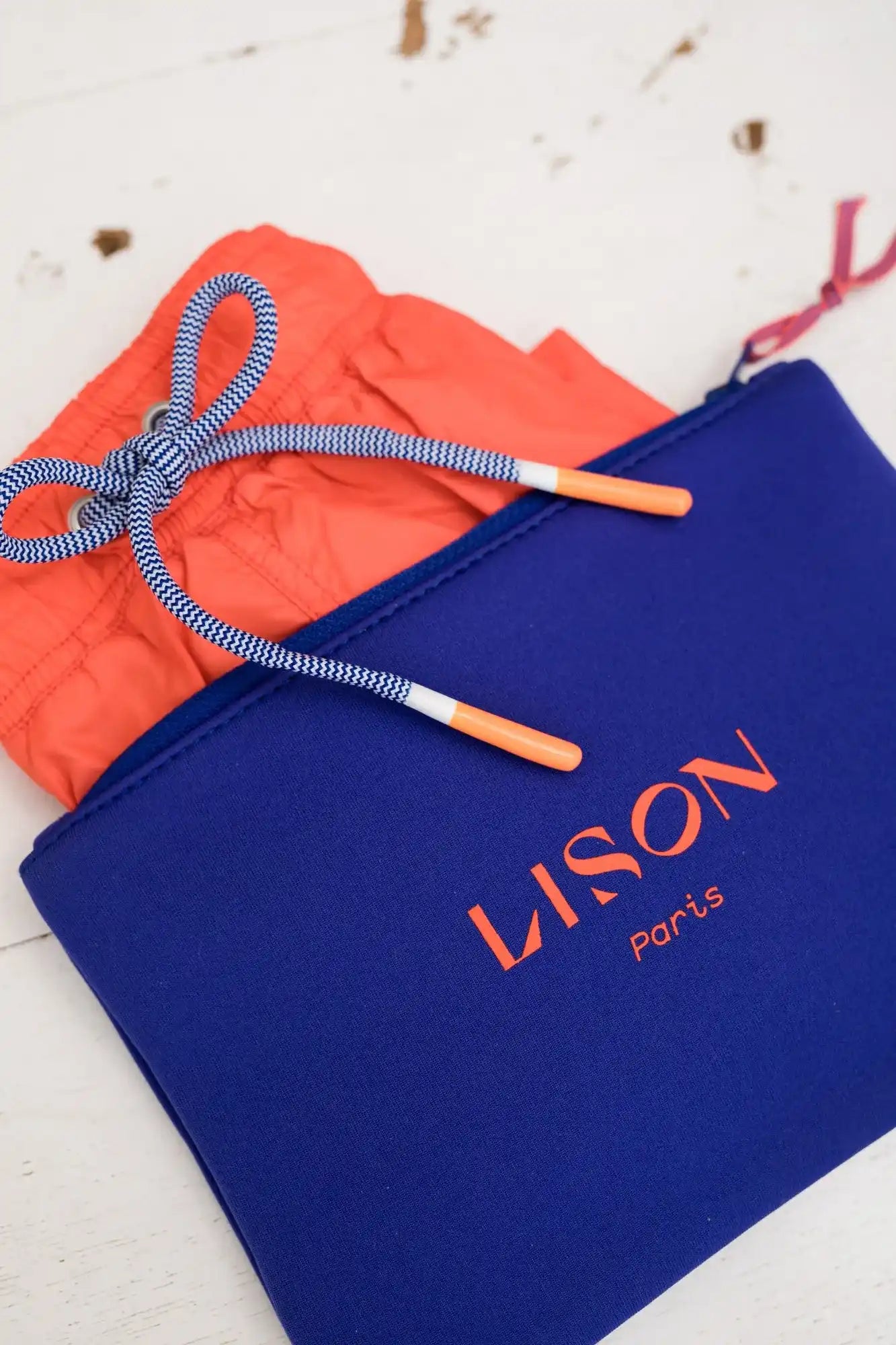 Bright orange boys' swim trunks with blue and white drawstring inside Lison Paris blue pouch.