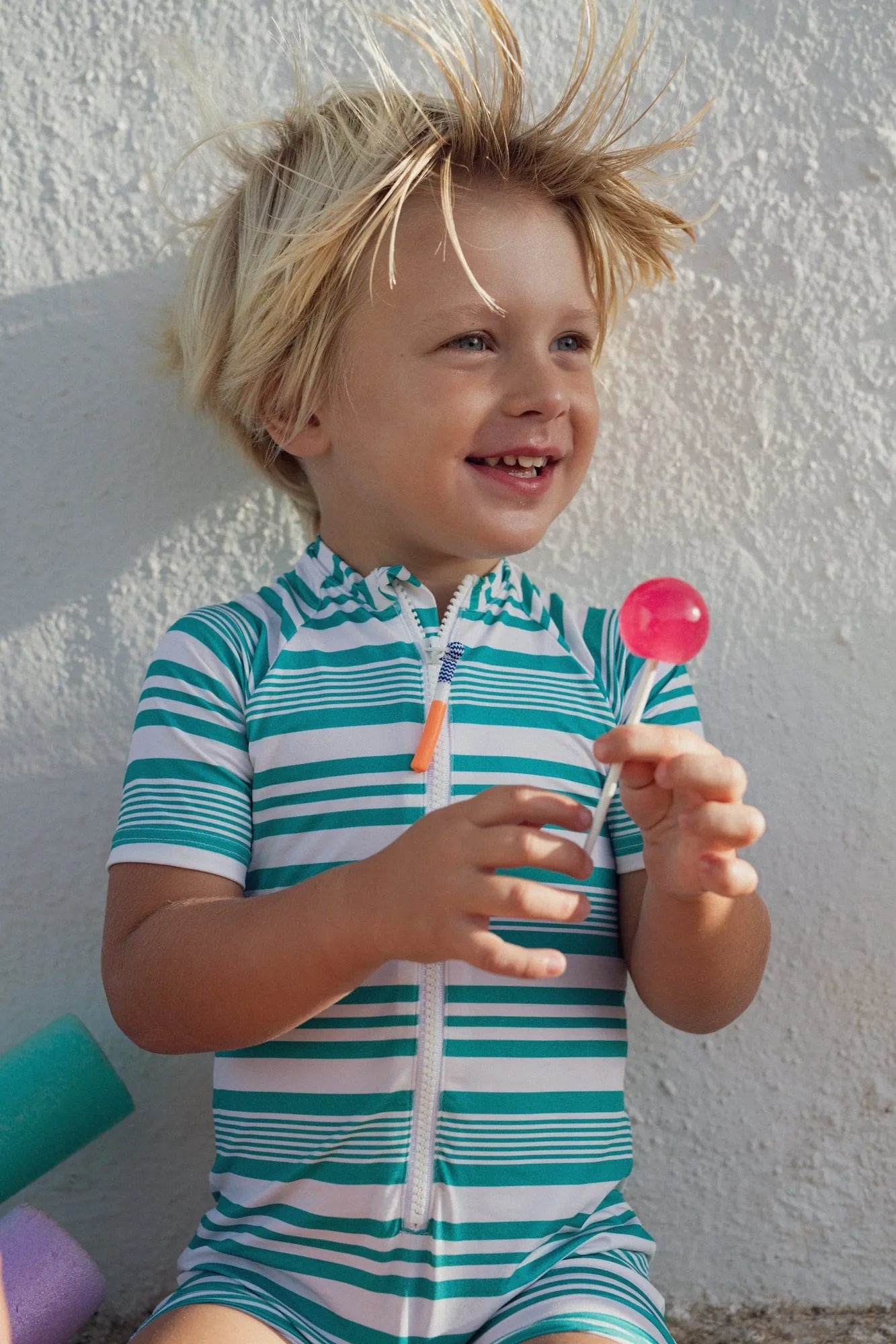 Smiling young boy in green and white striped short-sleeve UV protective swimsuit holding a pink lollipop